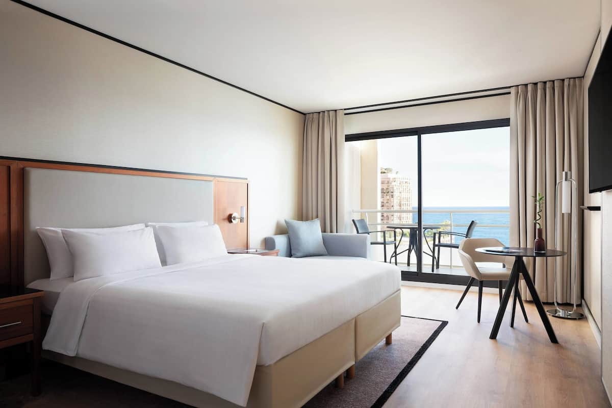 Une chambre d’hôtel avec un grand lit, un bureau avec une chaise, une petite table avec une chaise, et un balcon donnant sur la mer.