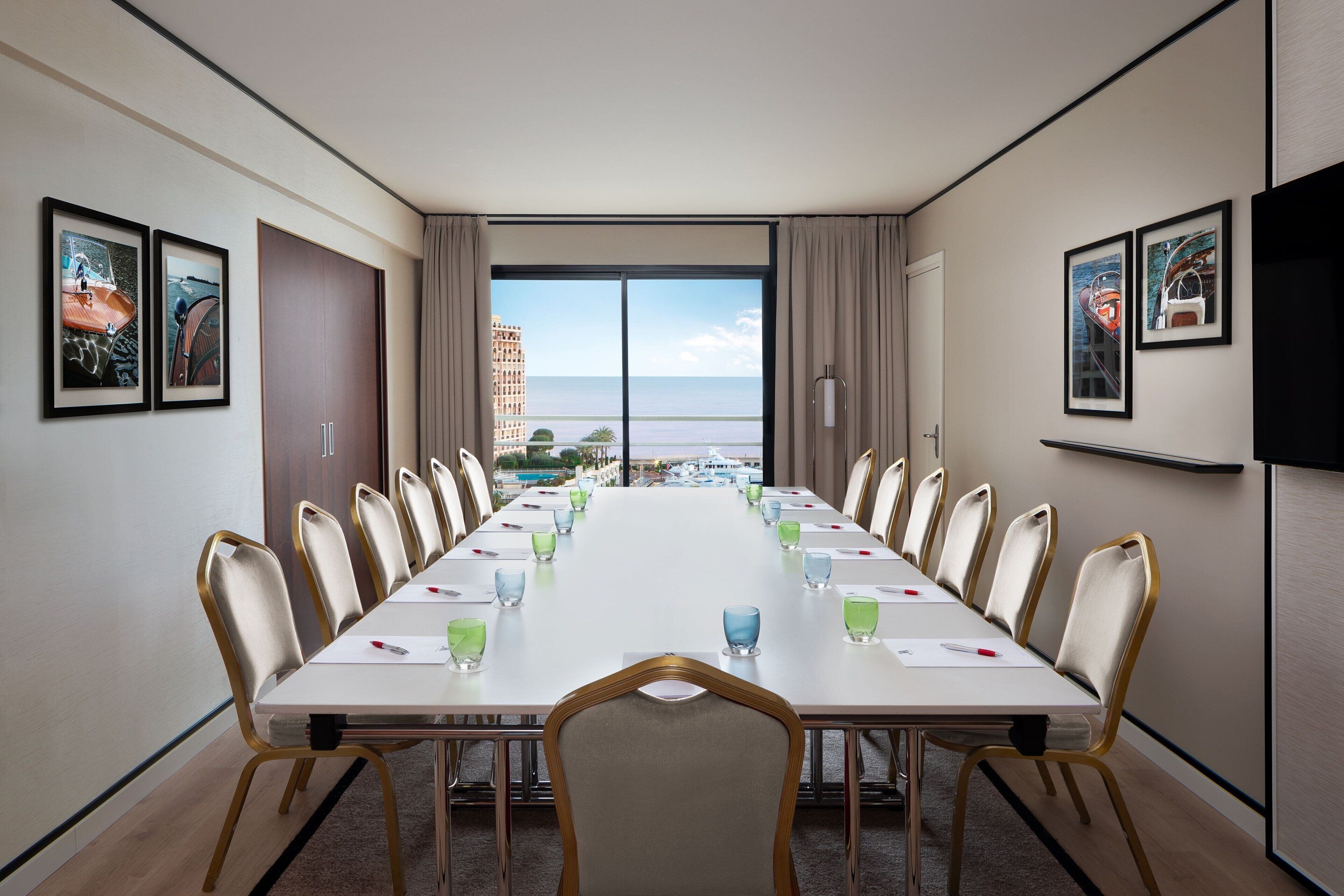 Un salón de conferencias con una larga mesa, sillas y un ventanal con vistas a edificios y al mar.