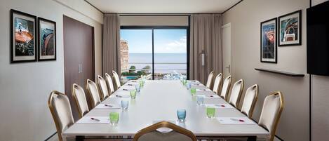Un salón de conferencias con una larga mesa, sillas y un ventanal con vistas a edificios y al mar.