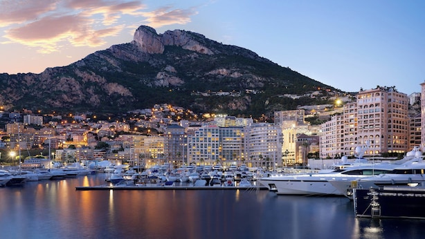 Un puerto deportivo con varios yates amarrados, un telón de fondo montañoso y un paisaje urbano con edificios iluminados.