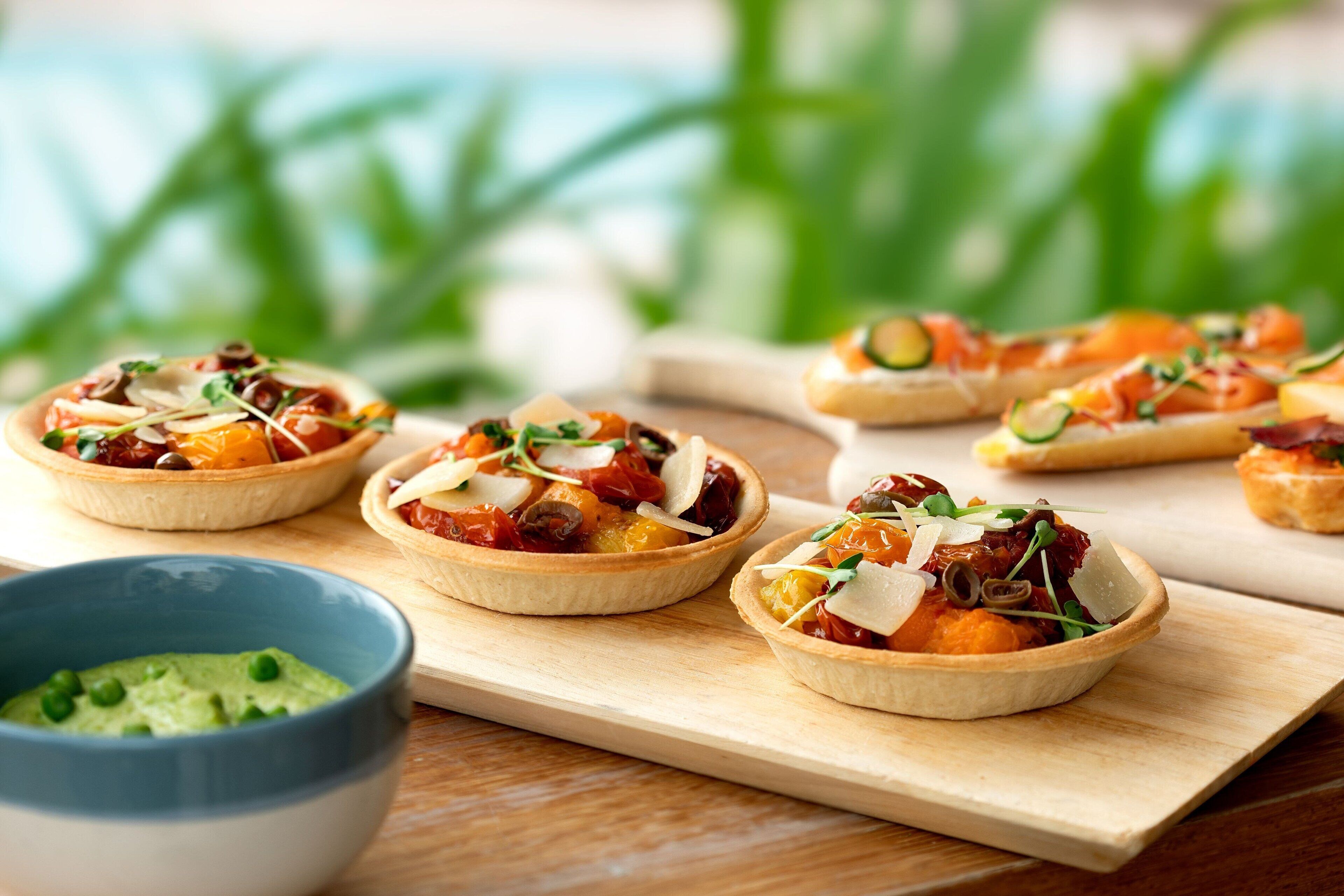Ein Holzbrett mit mehreren kleinen Tartlets, belegt mit verschiedenen Zutaten, einer Schüssel grüner Dip und einem unscharfen grünen Hintergrund.