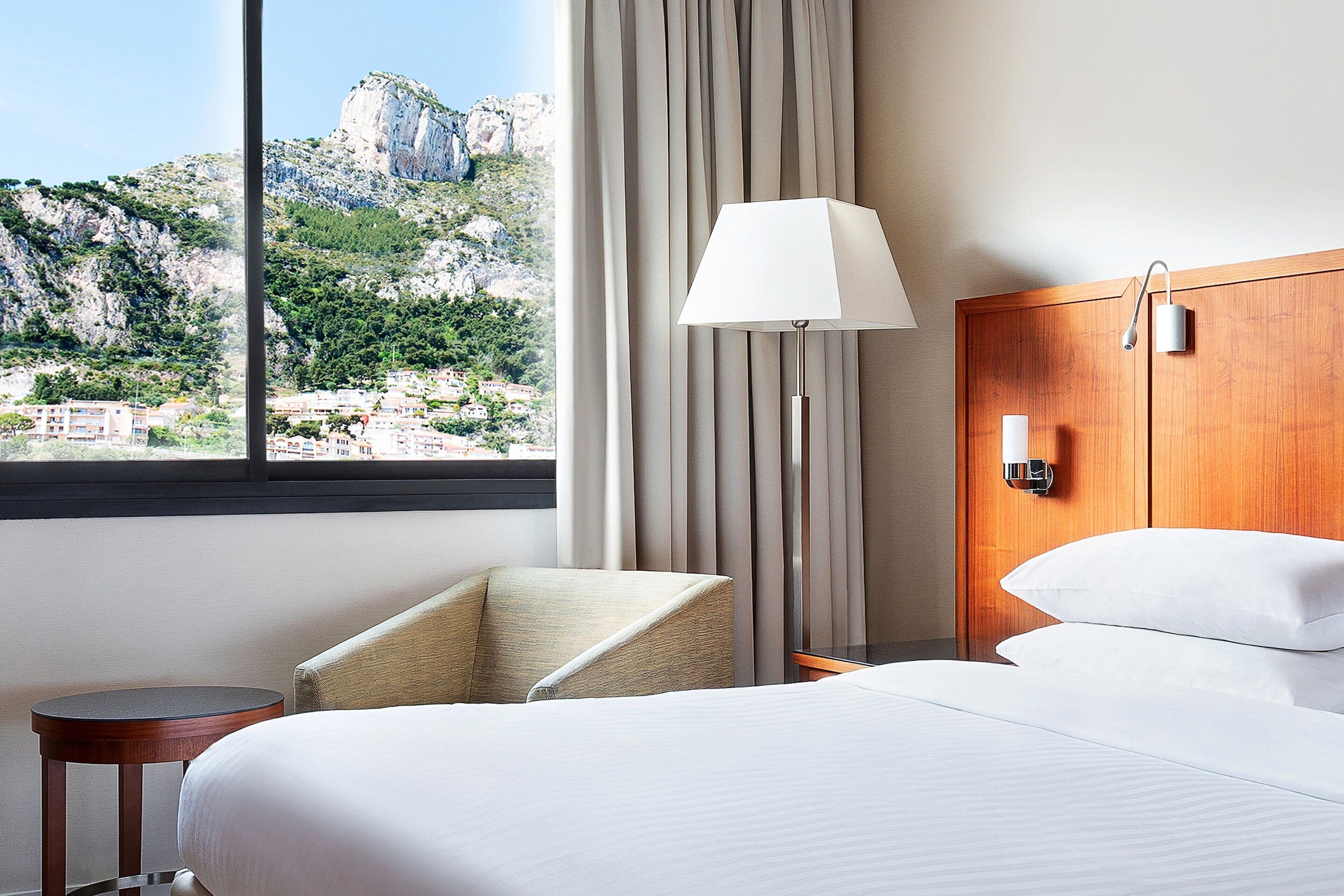 A hotel room with a bed, a chair, a lamp, and a view of a mountainous landscape.