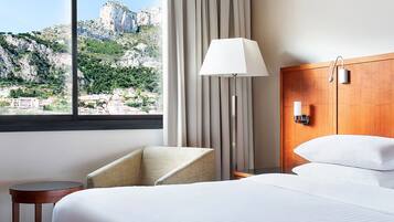 Habitación de hotel con cama, una silla, una lámpara y vistas a un paisaje montañoso.