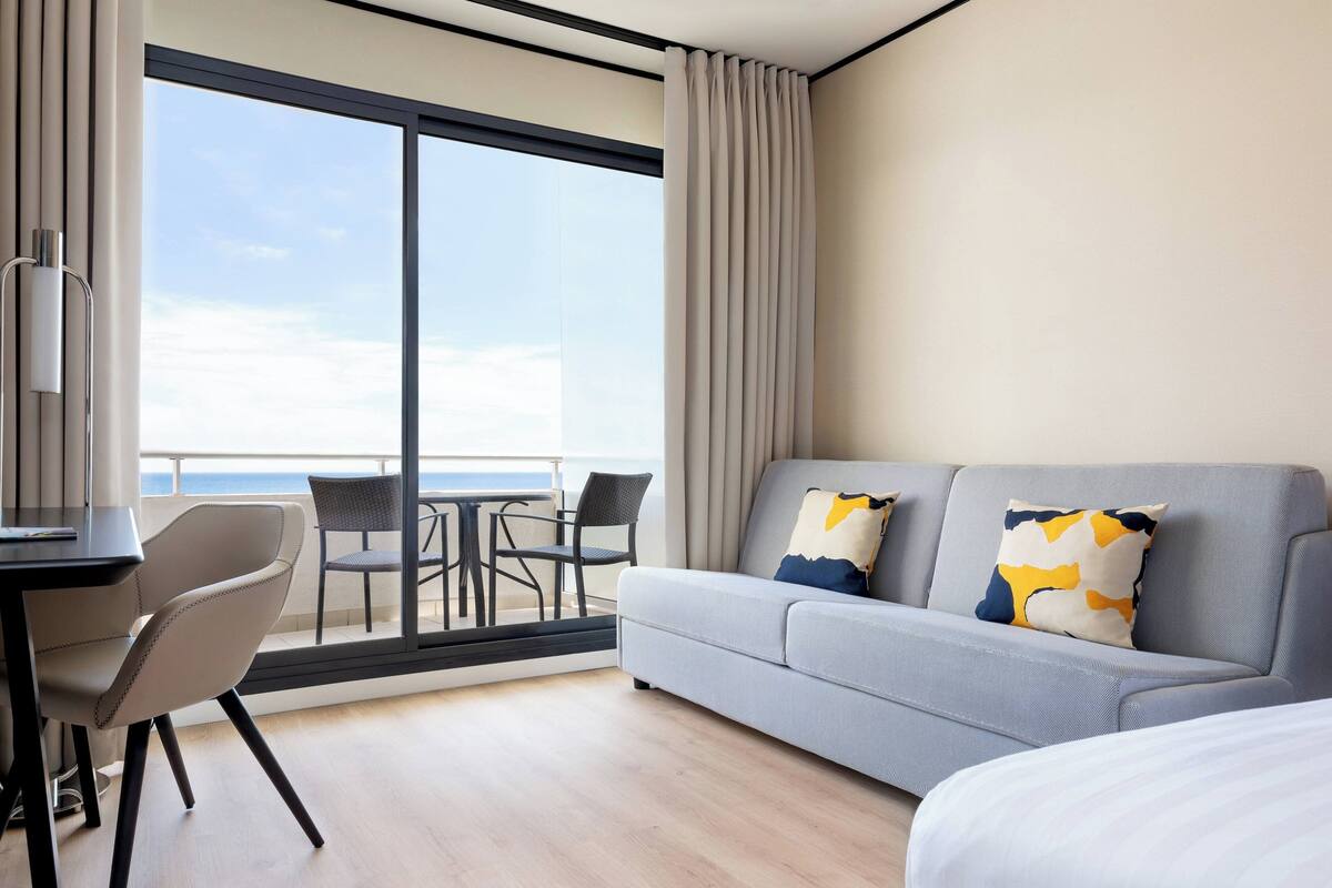Une chambre d’hôtel moderne dotée d’une grande porte-fenêtre coulissante donnant sur le balcon, d’un canapé gris agrémenté de coussins décoratifs, d’une petite table avec des chaises et d’un lit avec une literie blanche.