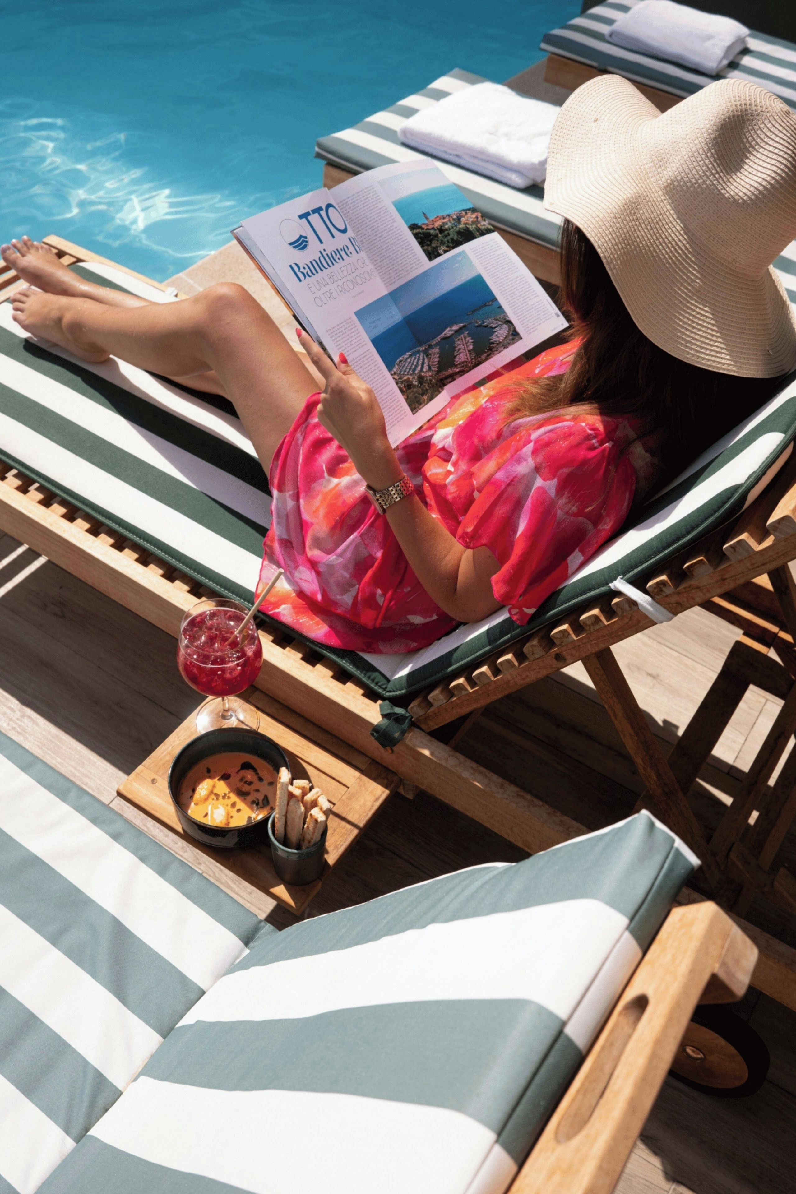A person lying on a striped lounge chair by a pool, reading a magazine.
