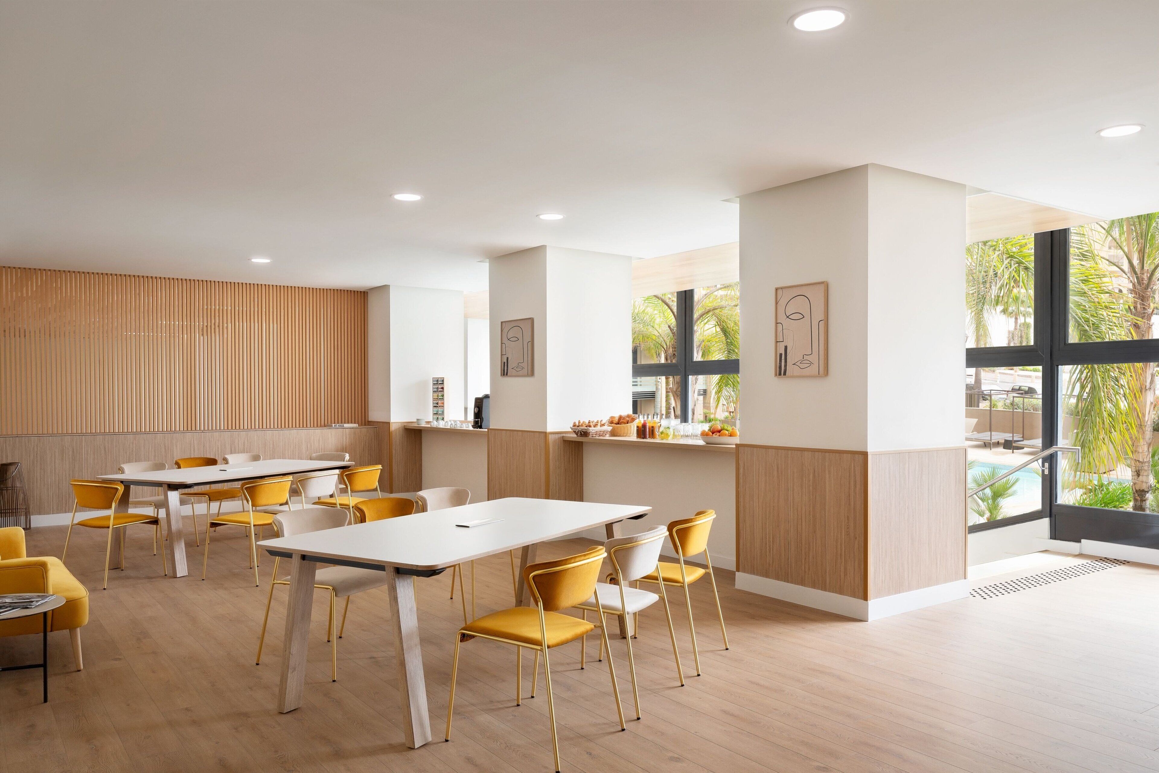 A modern dining area with a long table and yellow chairs, wooden paneling on the walls, and large windows offering a view of the outdoors.