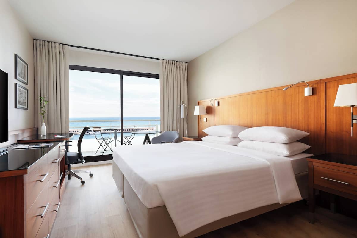 Une chambre d’hôtel avec un grand lit, un bureau avec un ordinateur, une chaise et une vue sur la mer par une grande fenêtre.