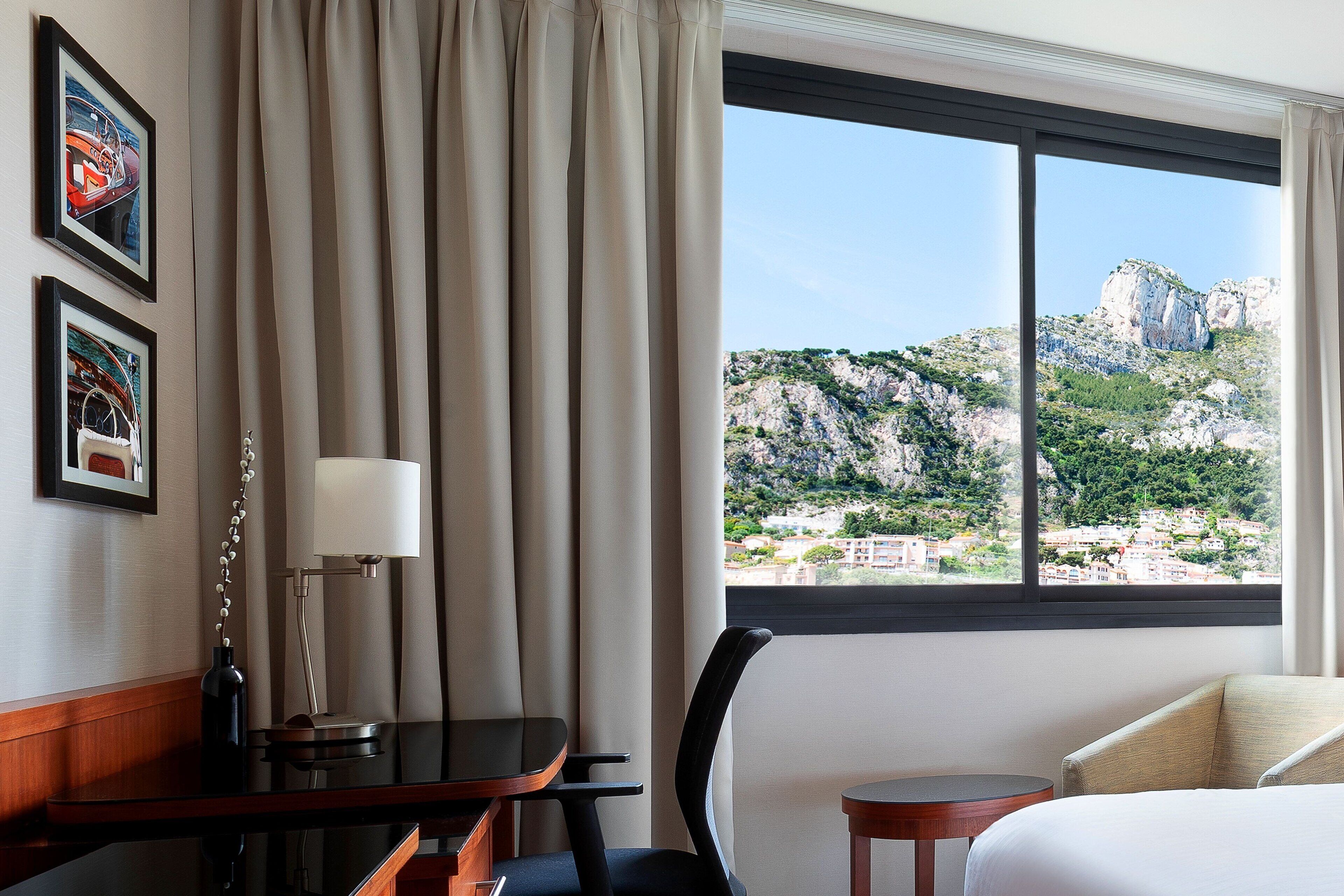 A hotel room with a bed, a desk, a chair, a lamp, and a view of a mountainous landscape through a large window.