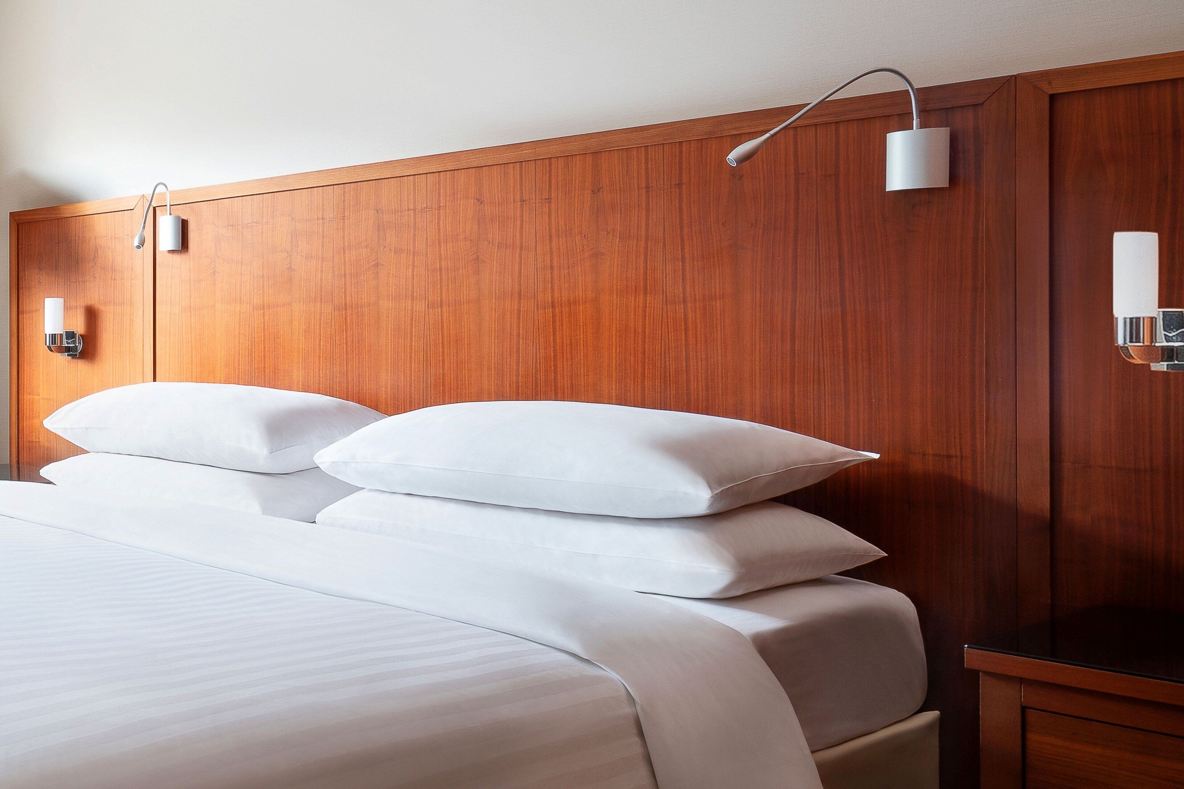 A neatly made bed with white linens and pillows, a wooden headboard, and two wall-mounted bedside lamps.