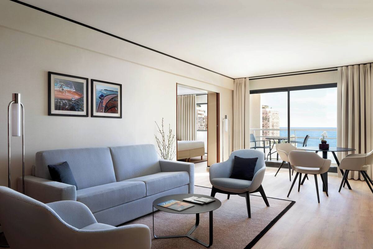 Un salon moderne avec un canapé, des fauteuils, une table basse et un coin repas comprenant des chaises et une table. Deux tableaux encadrés ornent le mur, ainsi qu’une lampe sur pied. De grandes fenêtres offrent une vue sur la mer.