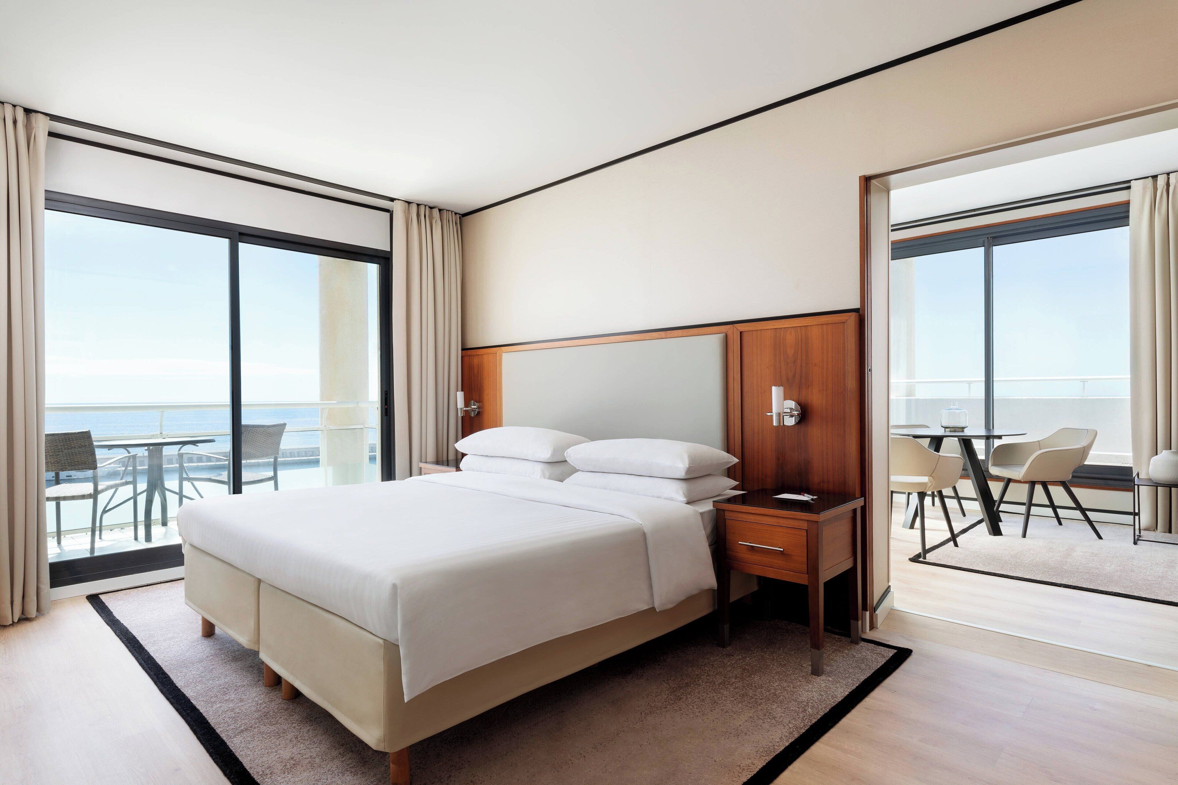 A modern hotel room with a large bed, a wooden nightstand, a desk, and a chair. There is a balcony with a table and chairs, and a view of the ocean.
