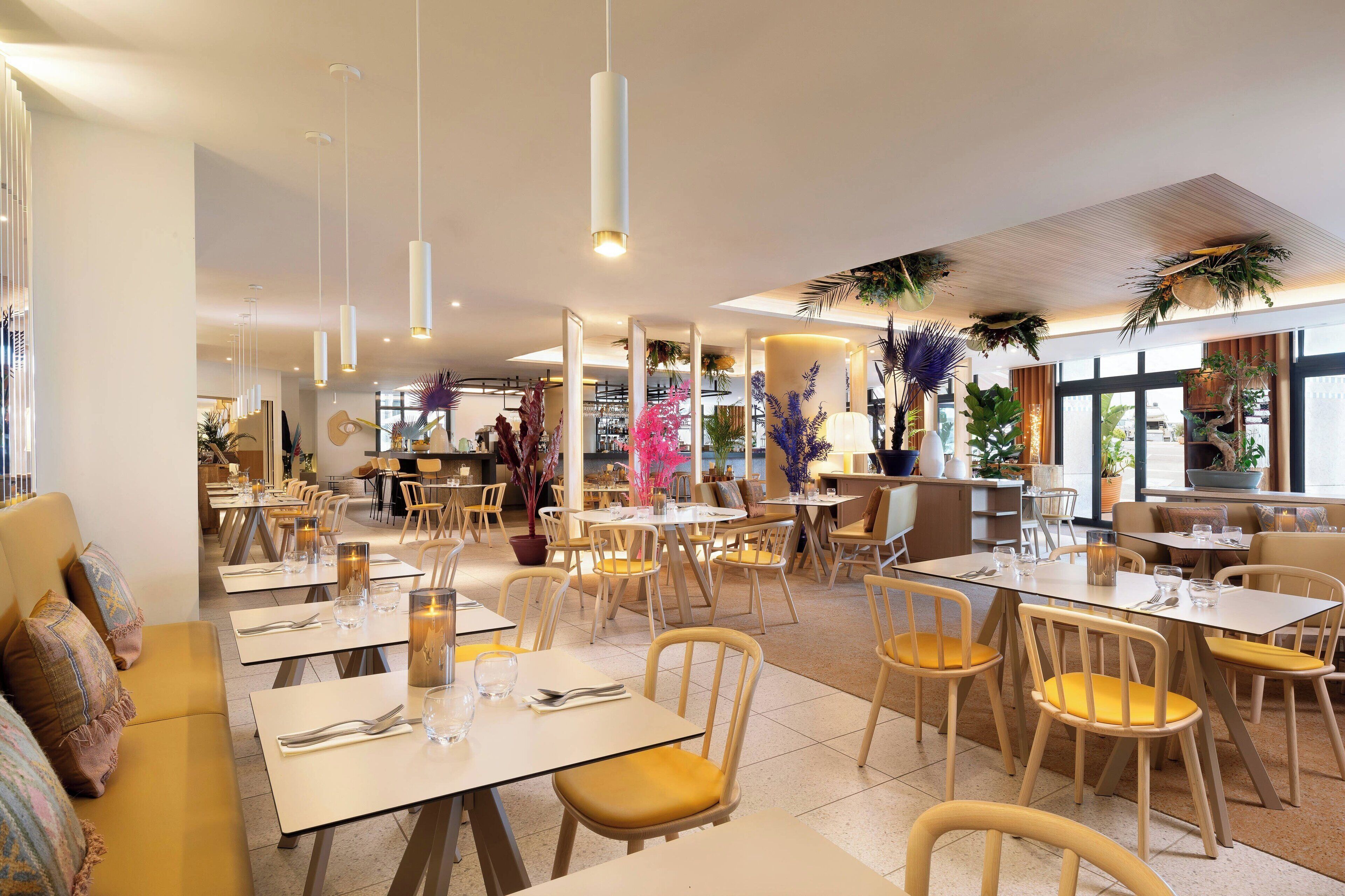 A restaurant with multiple tables and chairs, indoor plants, and a modern ceiling design.