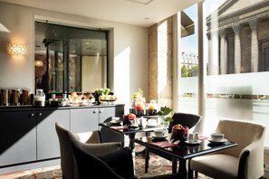 A modern hotel room with a dining area featuring a table set for breakfast, a large window with a view of a historic building, and a mirror reflecting part of the room. - Madeleine Plaza (Paris)