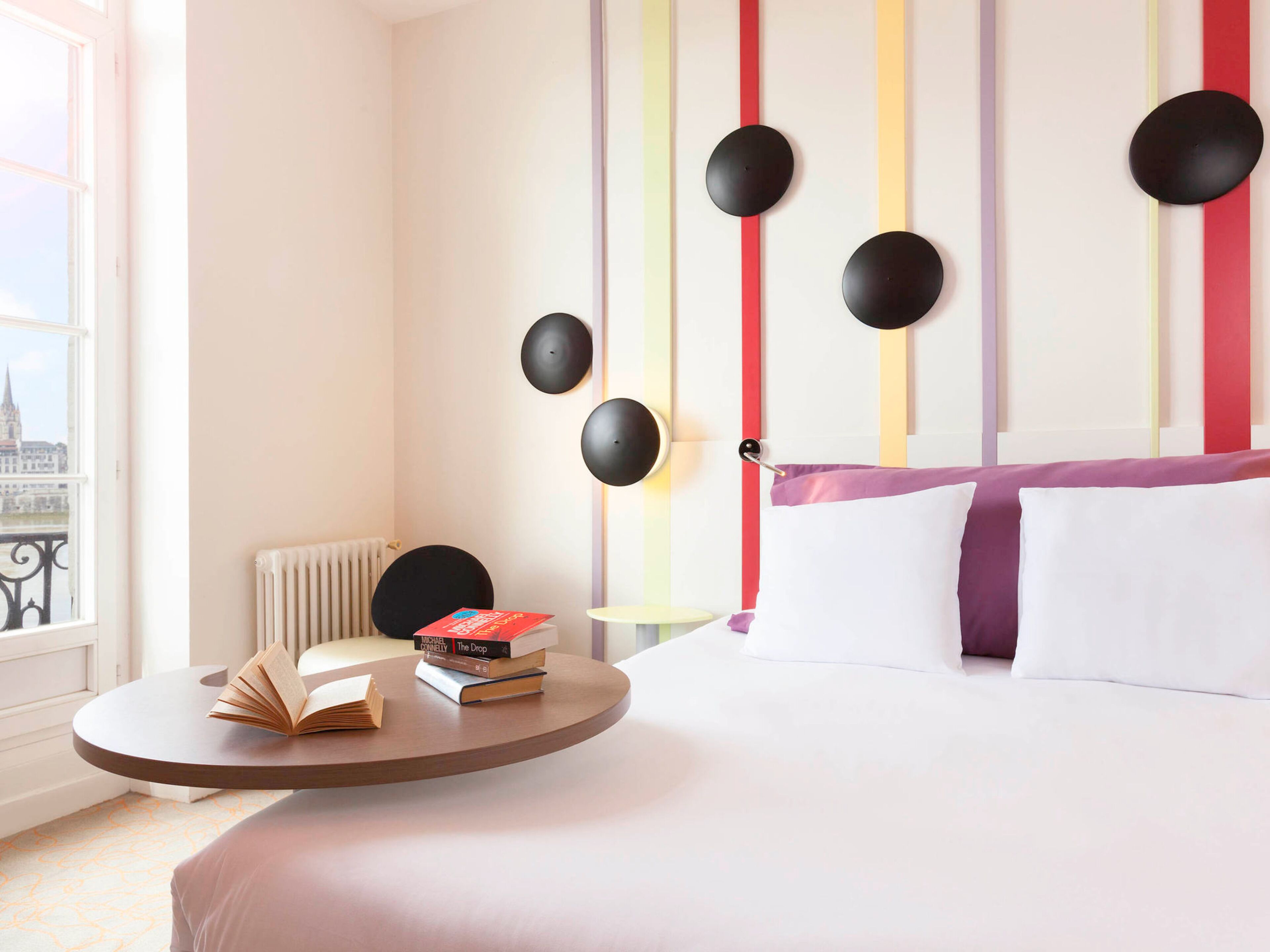A hotel room with a bed, a round table with books, a radiator, and a view of a cityscape through a window.
