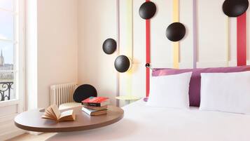 A hotel room with a bed, a round table with books, a radiator, and a view of a cityscape through a window.