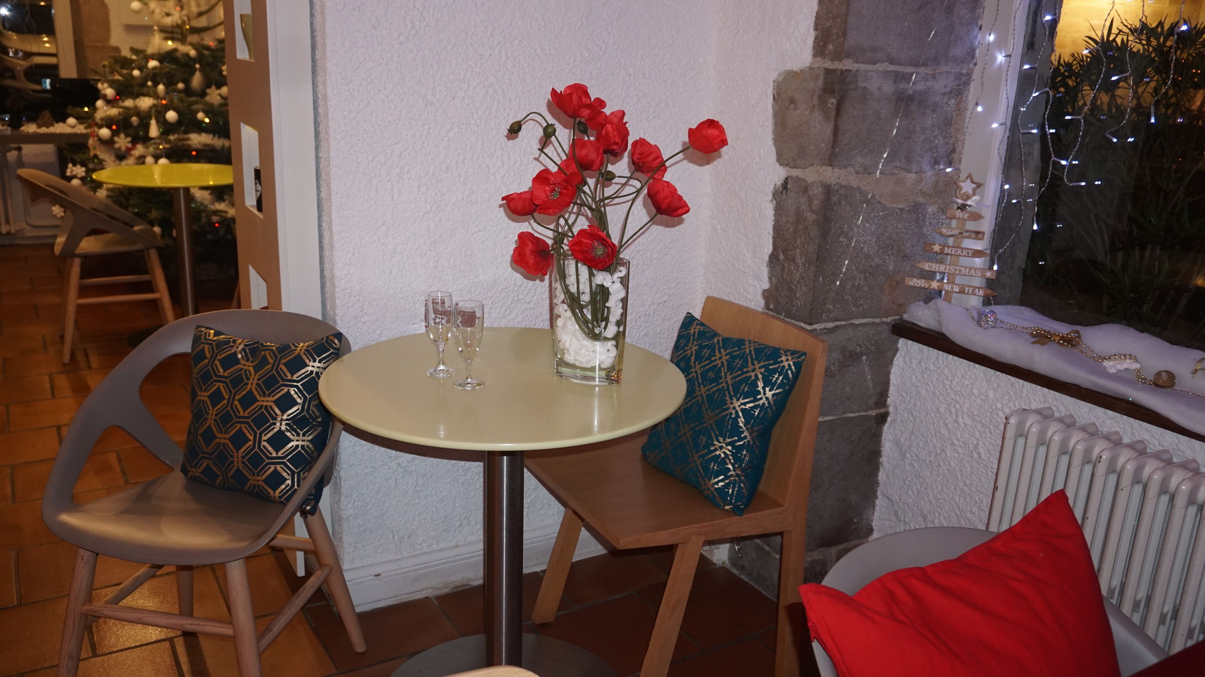 Un coin repas avec une table ronde, deux chaises, un vase avec des fleurs rouges et, au fond, un sapin de Noël.