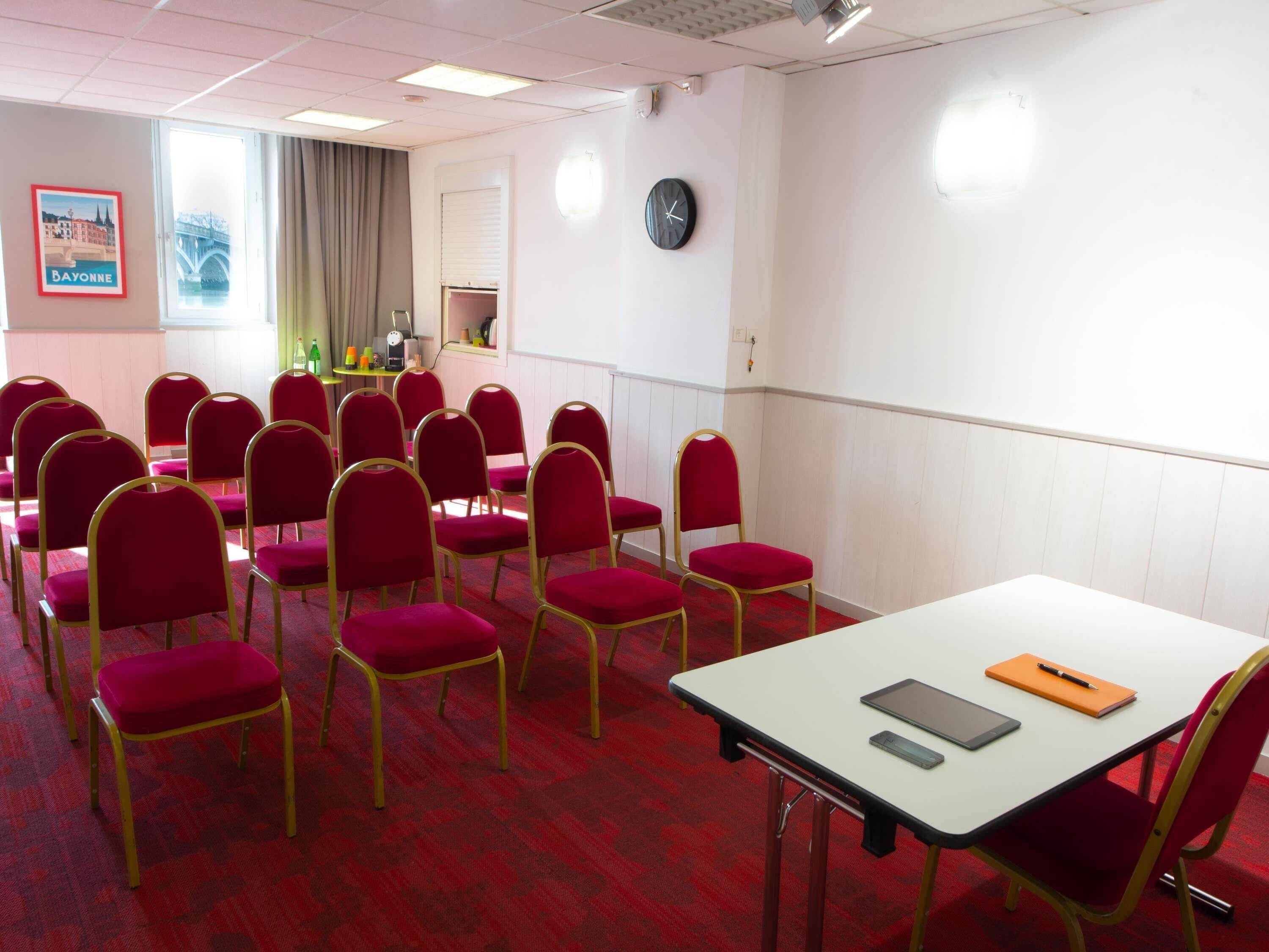 Une salle de conférence avec des chaises rouges, une table blanche, une horloge et une affiche représentant un pont.