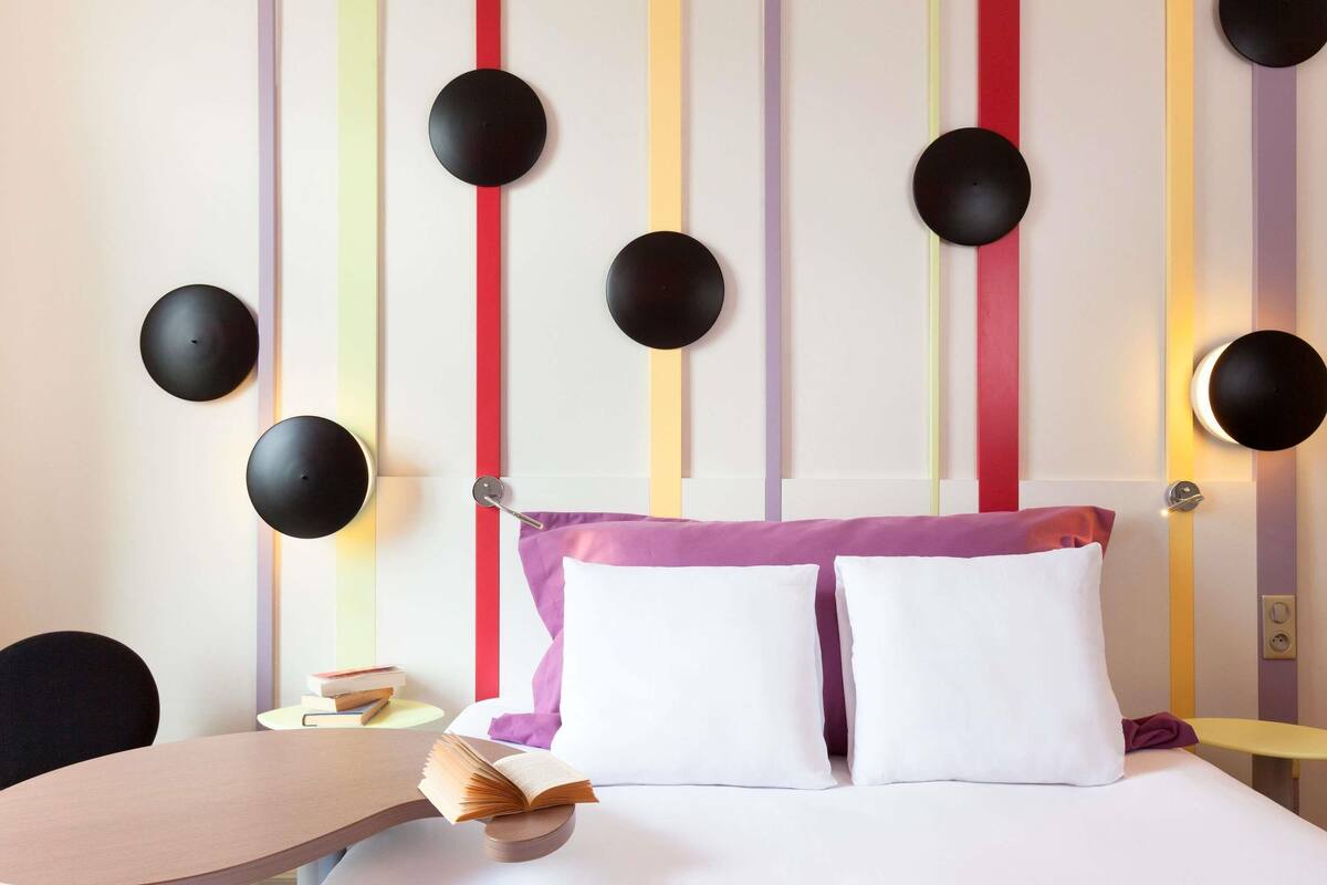 Una habitación de hotel con cama, mesitas de noche, una silla y una estantería.