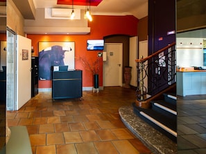 Un hall con mostrador de recepción, televisor, escalera y suelo de baldosas.