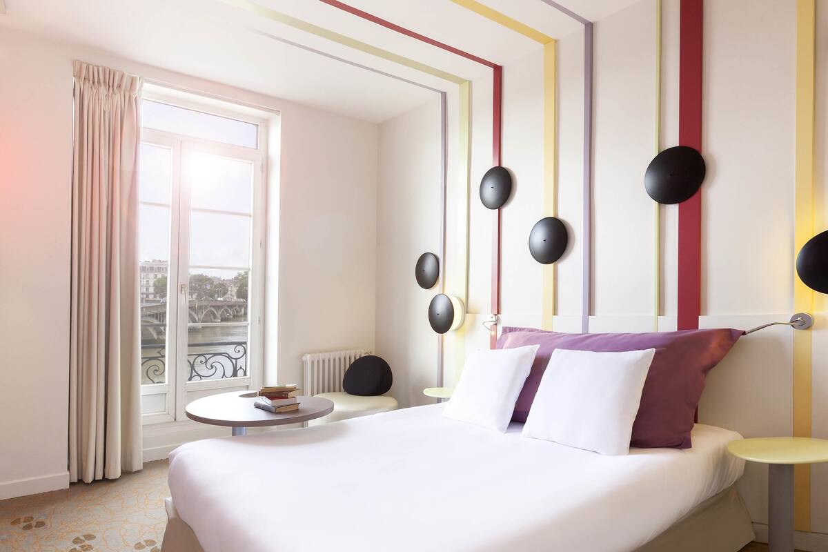 Habitación de hotel con cama, mesitas de noche, ventana con cortinas y vistas a un puente, además de un radiador.
