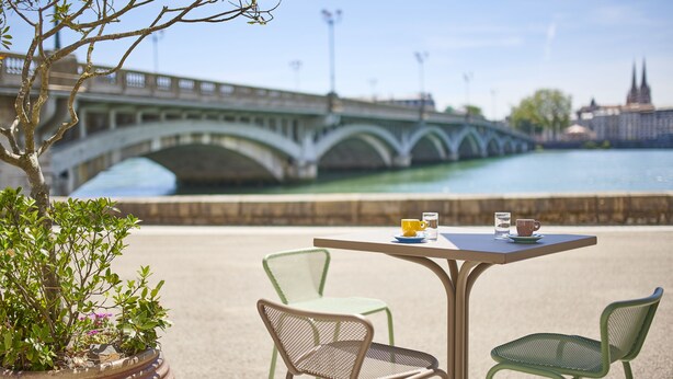 Une table avec des chaises et un pot de fleurs, donnant sur une rivière, avec un pont et des bâtiments en arrière-plan.