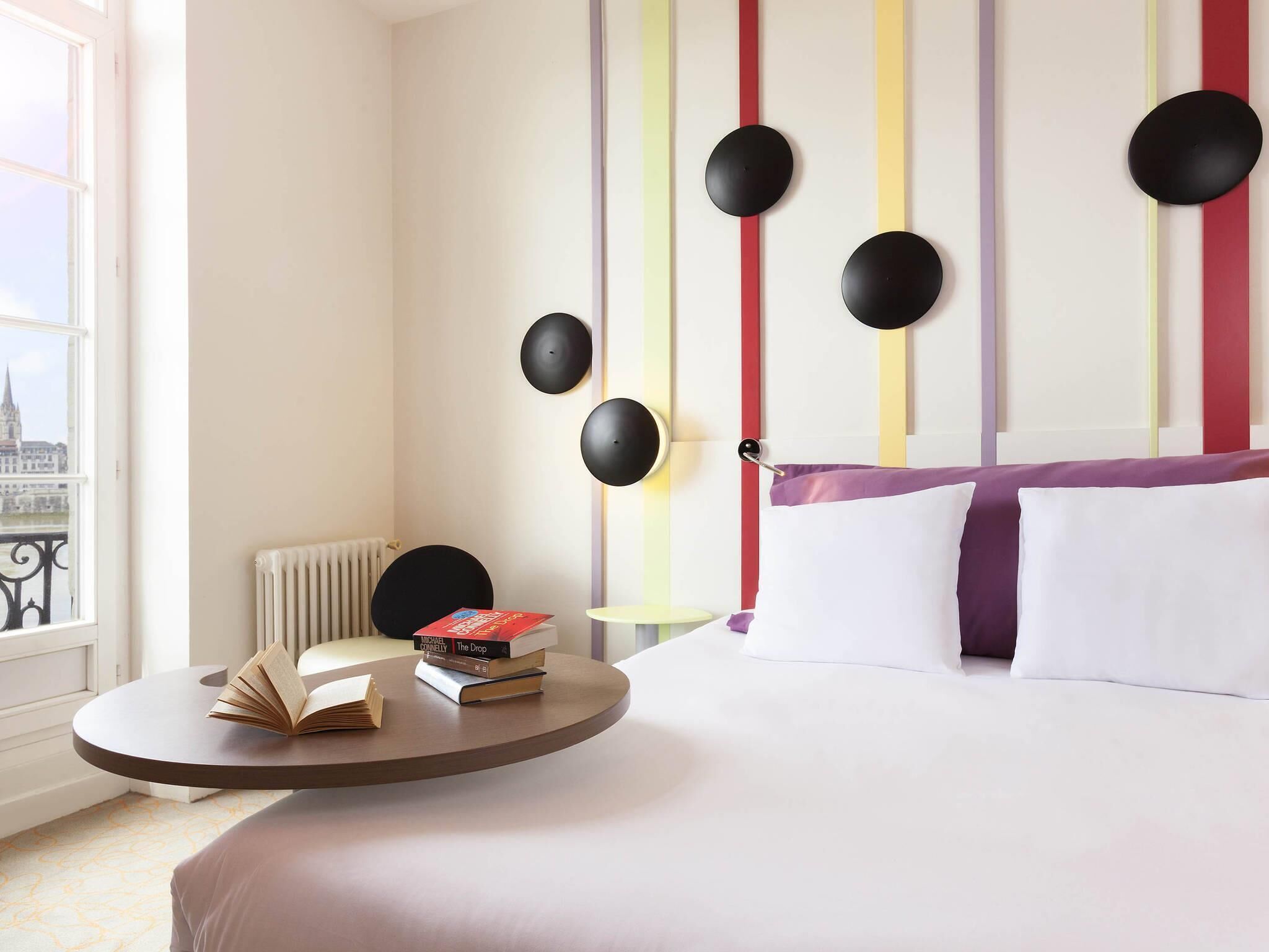 A hotel room with a bed, a round table with books, a radiator, and a view of a cityscape through a window.