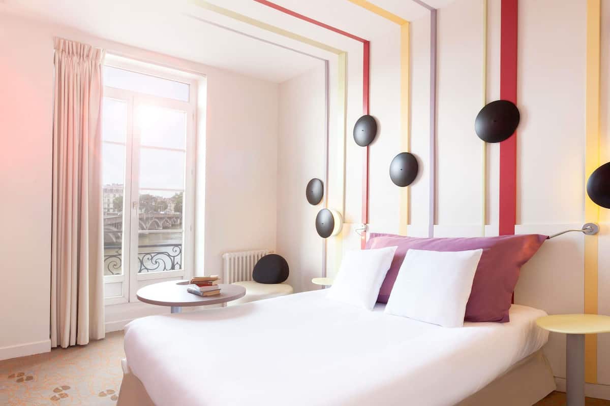 A hotel room with a bed, a round table, a window with curtains, and a view of a bridge.