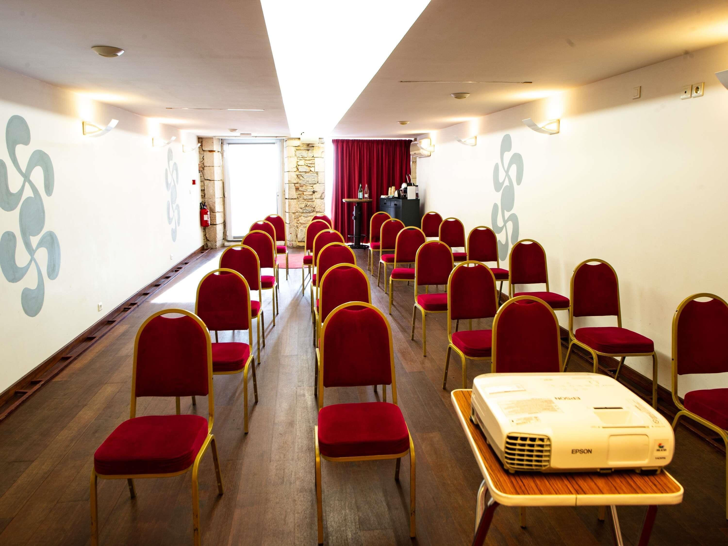 Uma sala de conferências com fileiras de cadeiras vermelhas, um projetor sobre uma mesa e uma parede de pedra com uma porta.