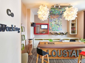 A modern co-working space with a large wooden sign reading 'CO WORKING LOUSTAU,' a television, a large decorative wall piece, and a bookshelf with mugs. - ibis Styles Bayonne Centre Gare (Bayonne)