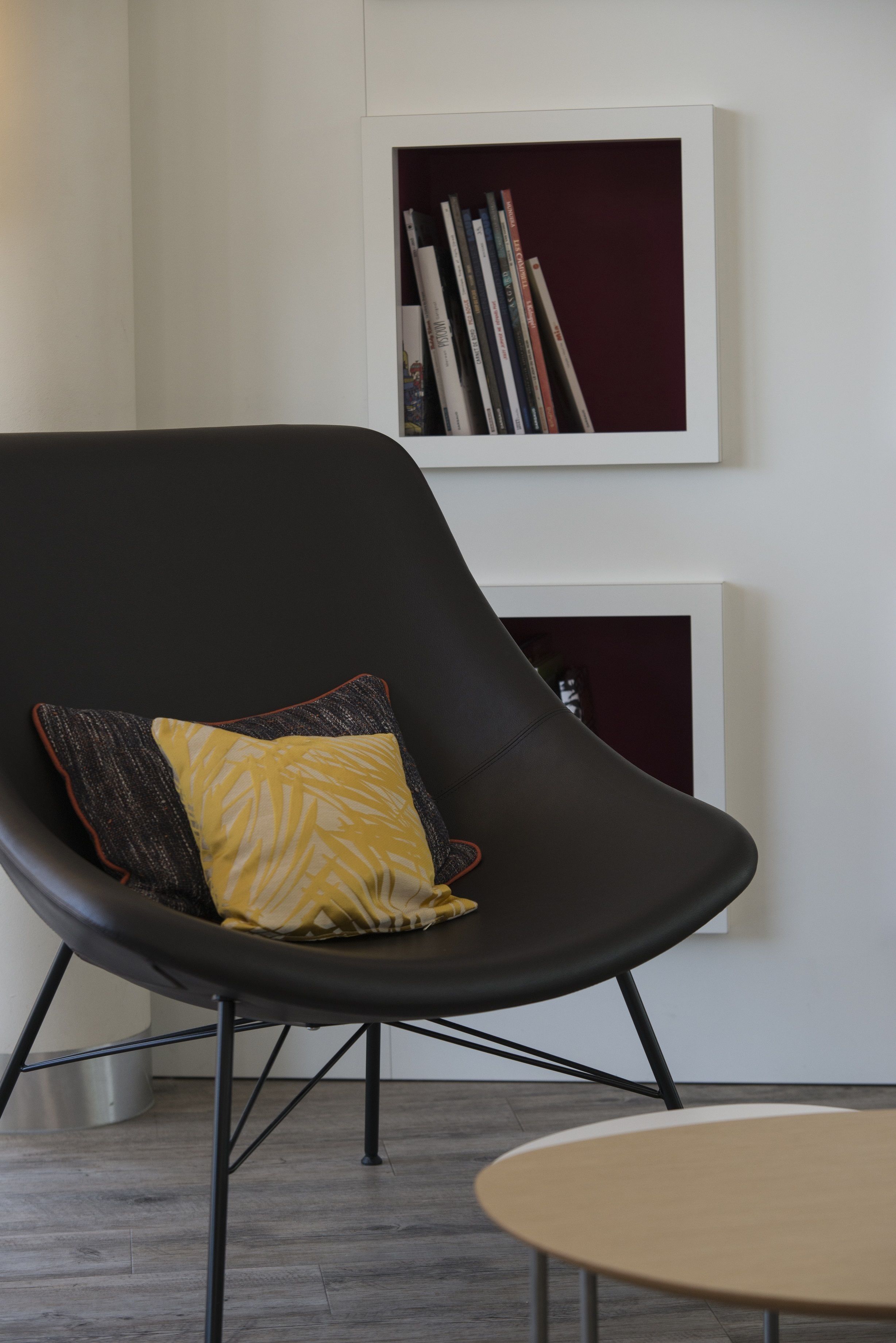Une pièce moderne avec une chaise noire, un coussin jaune, une petite table ronde et une bibliothèque adossée au mur.