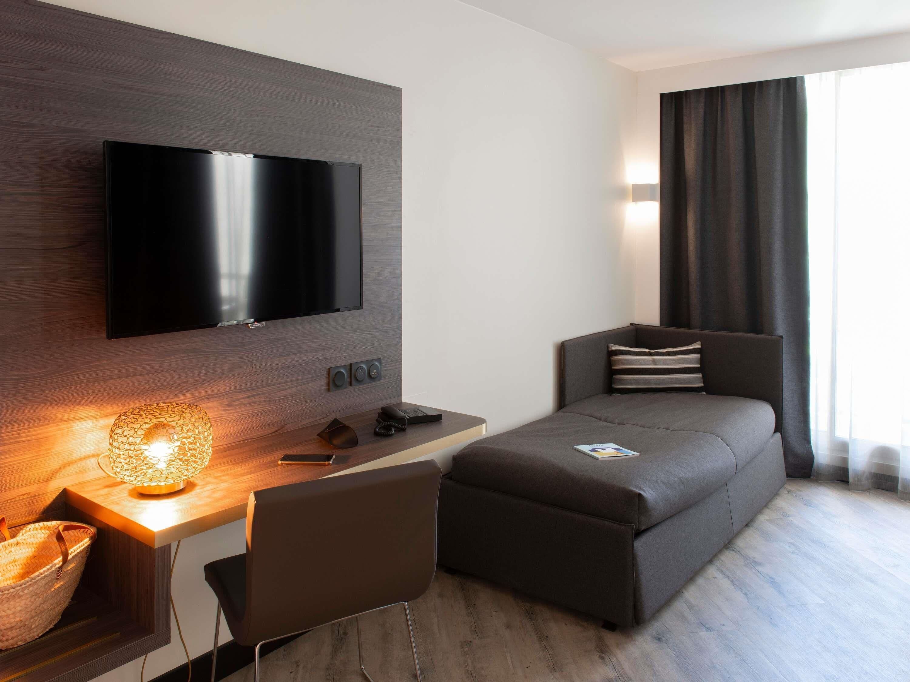 A modern hotel room with a flat-screen TV, a desk with a lamp, a sofa, and a window with curtains.