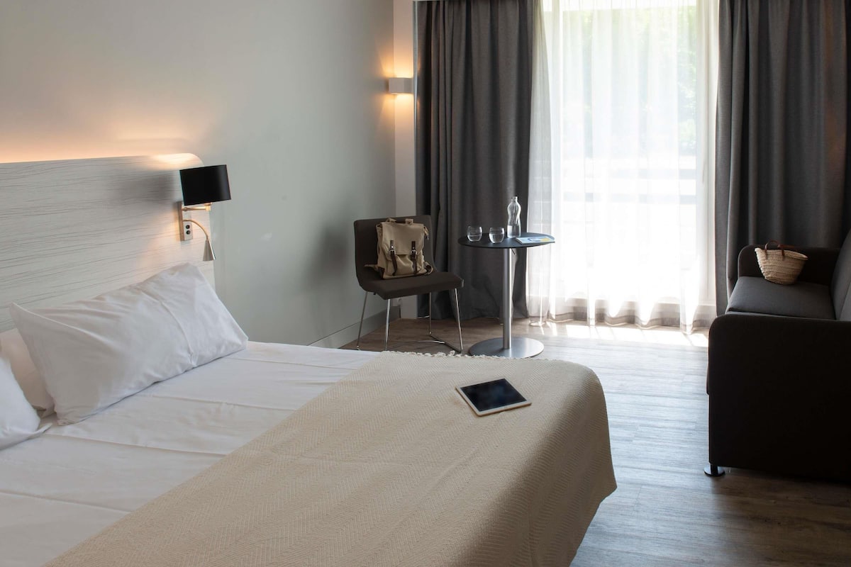 A hotel room with a bed, a desk, a chair, a sofa, and a window with curtains.