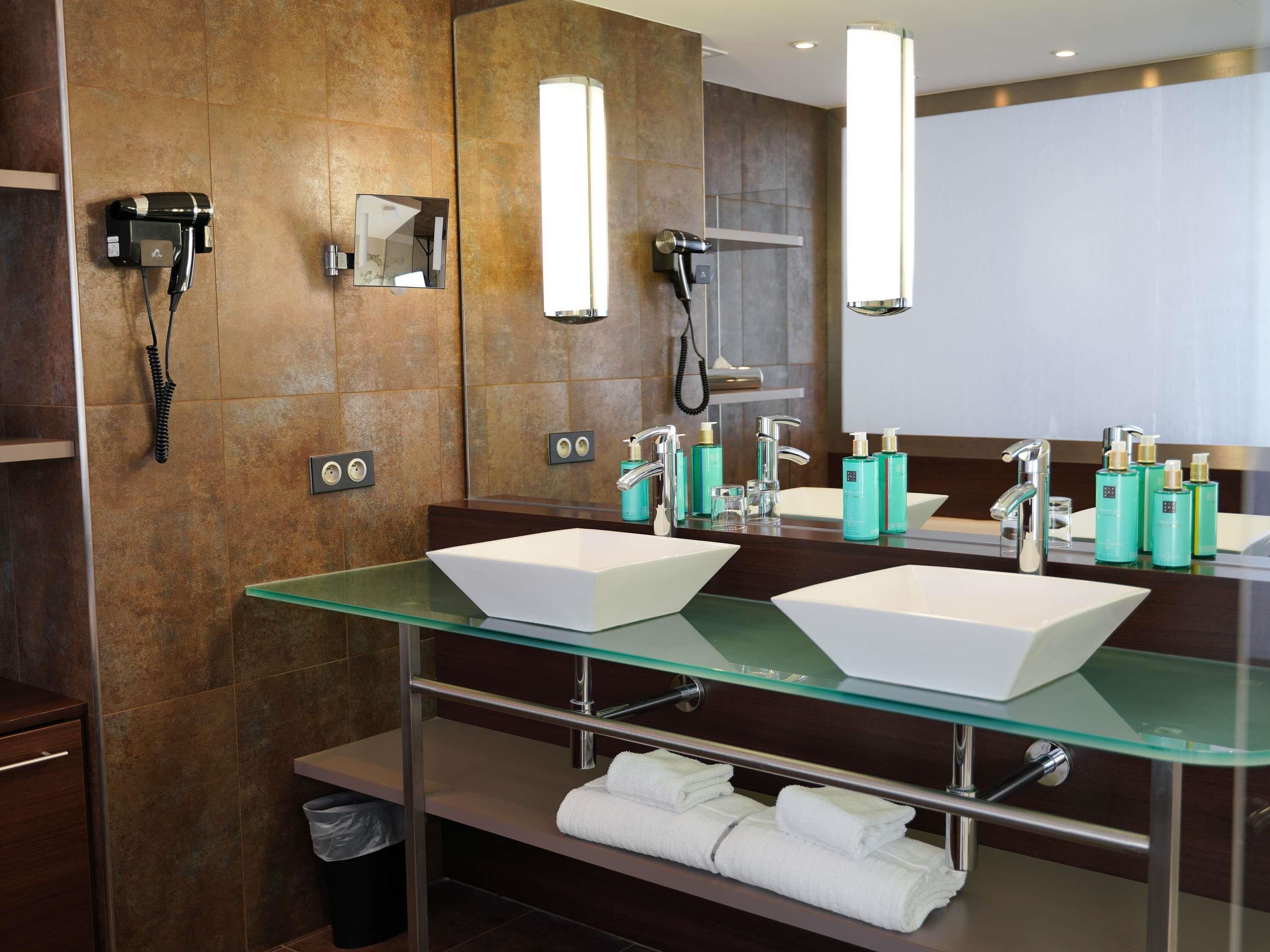 Un baño moderno con dos lavamanos, un secador de pelo, un espejo y una repisa de vidrio con toallas.