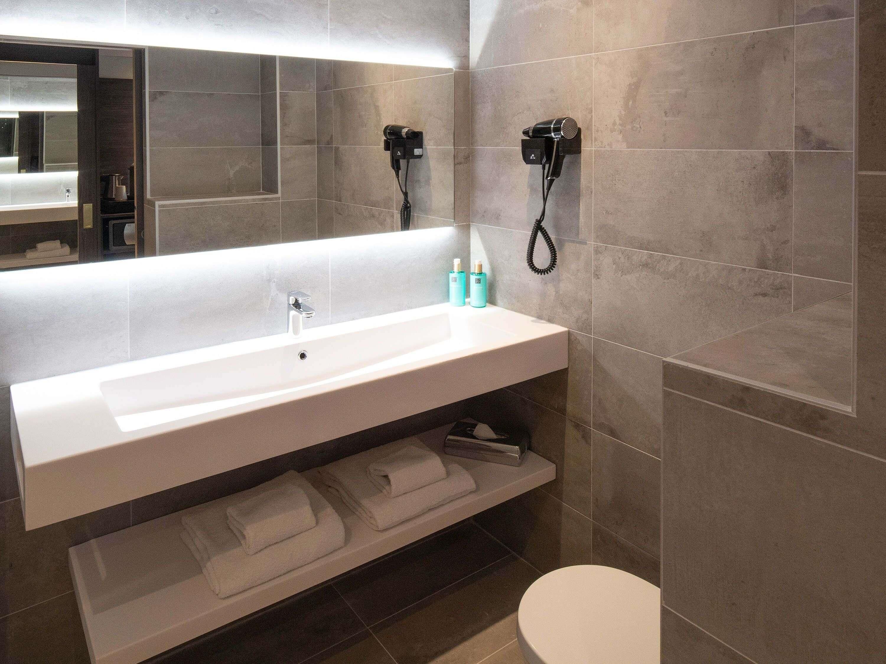 A modern bathroom with a large sink, two hair dryers mounted on the wall, and a toilet.