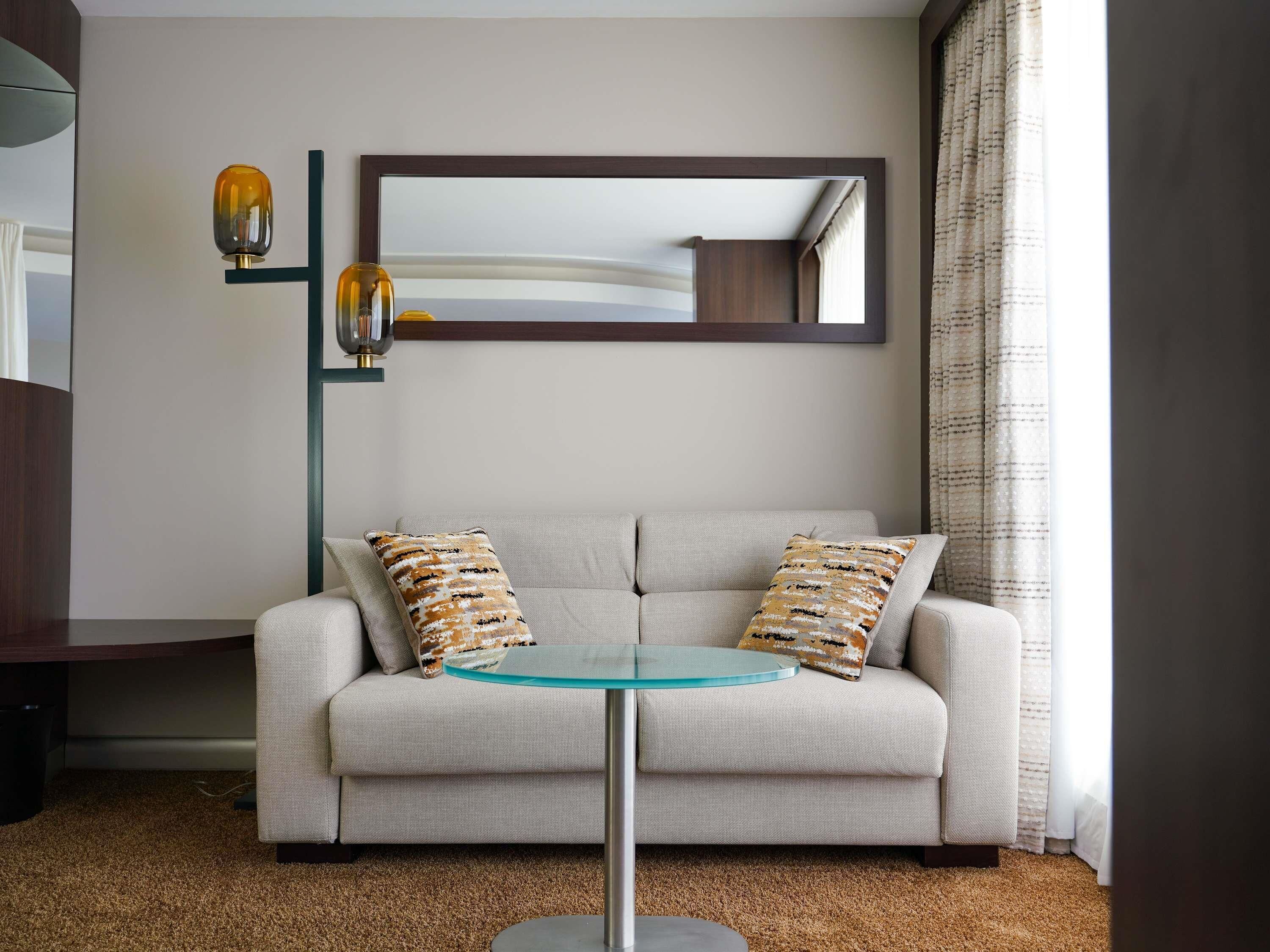 A modern living room with a sofa, two patterned pillows, a glass coffee table, a floor lamp, and a mirror on the wall.