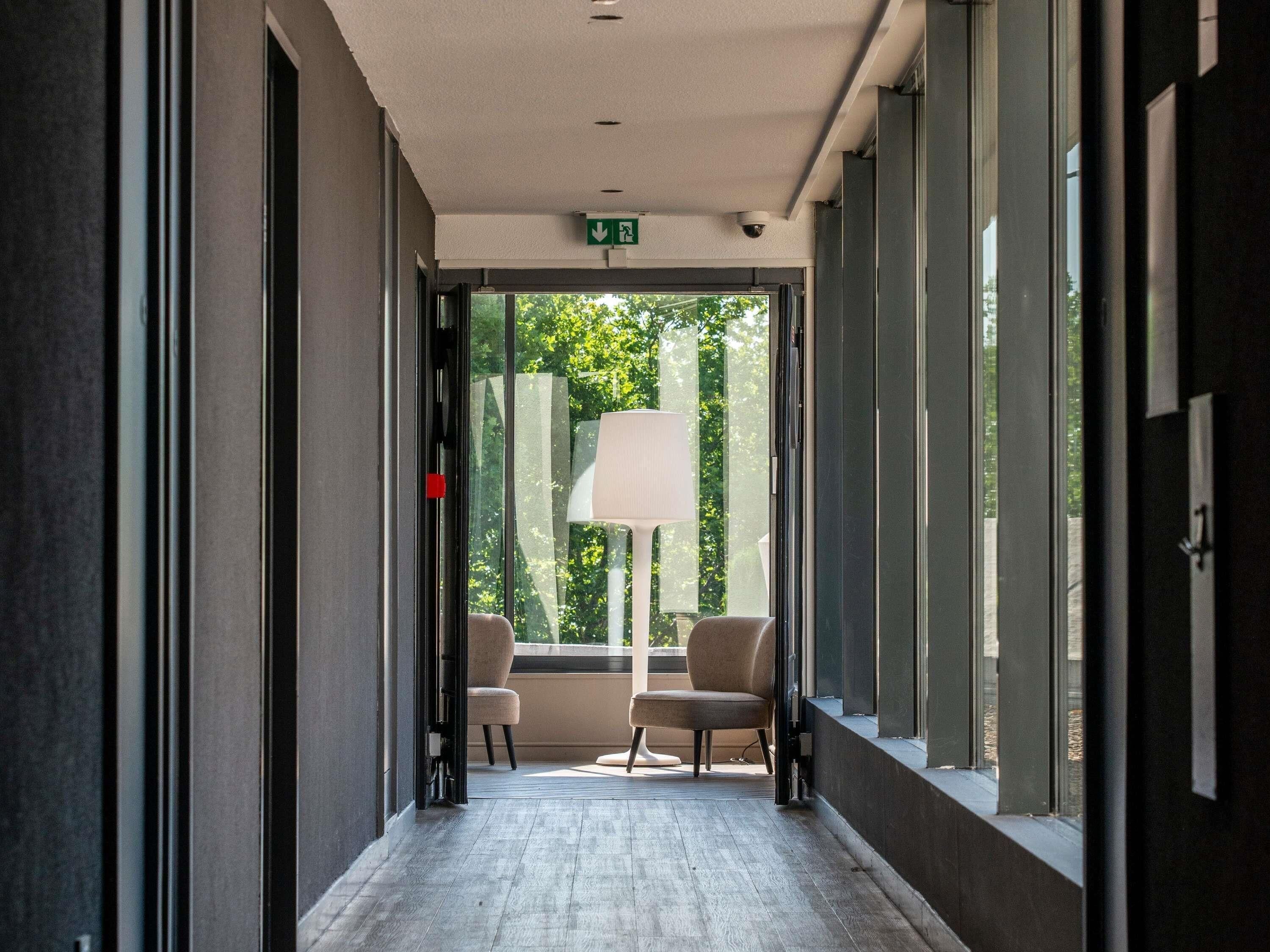 Ein Korridor mit Deckenleuchte, einem Fluchtzeichen, zwei Stühlen und einer Stehlampe.