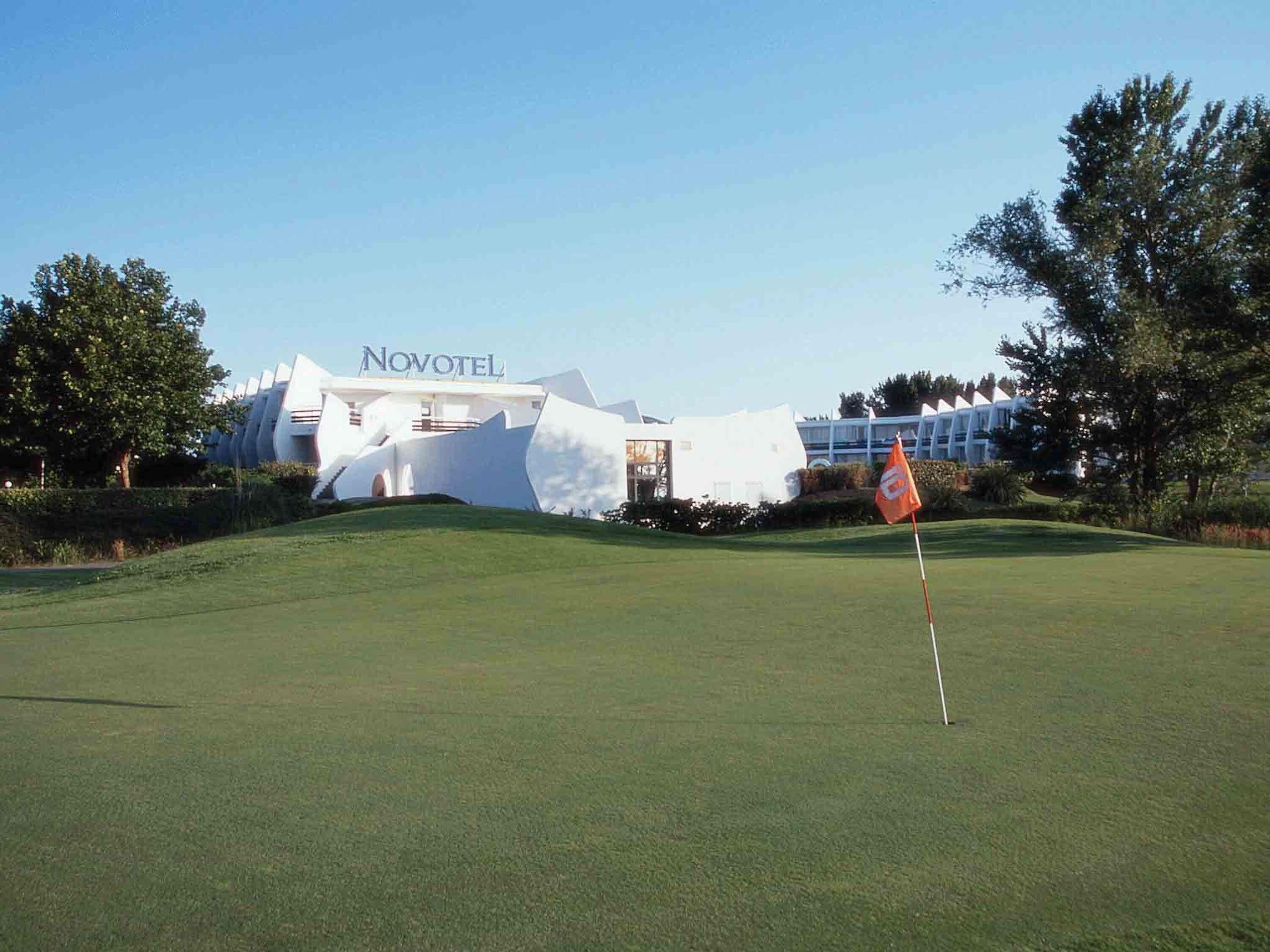 A golf course with a flag, a building labeled 'Novotel,' and surrounding greenery.