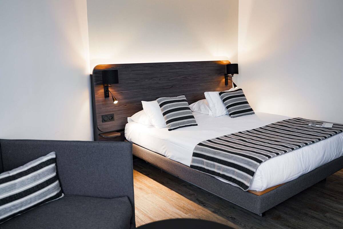 Ein modernes Schlafzimmer mit einem Bett, Nachttischlampen, einem grauen Sofa und Holzboden.