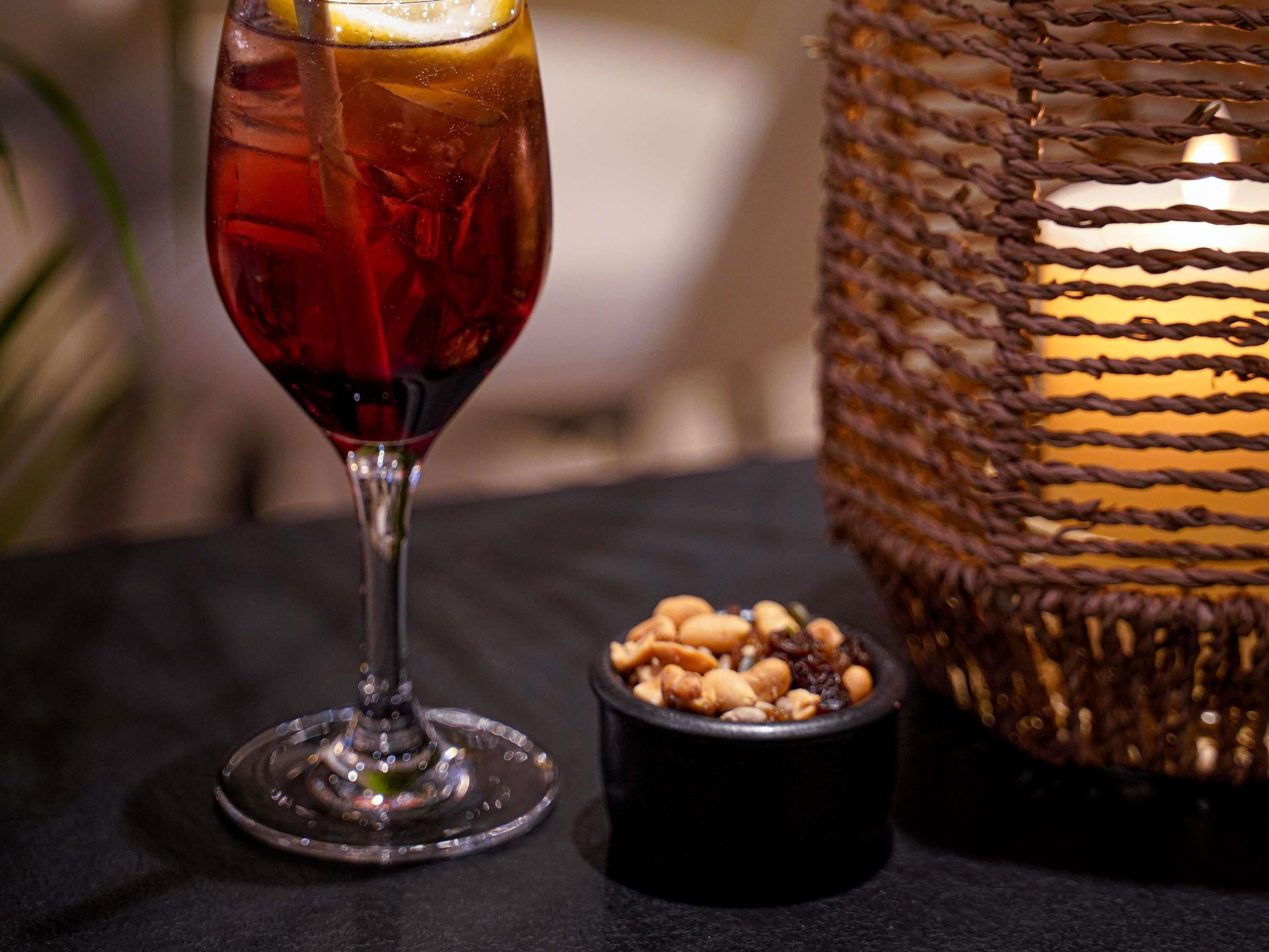 A glass of red beverage with a lemon slice, a small bowl of nuts, and a woven basket with a candle.
