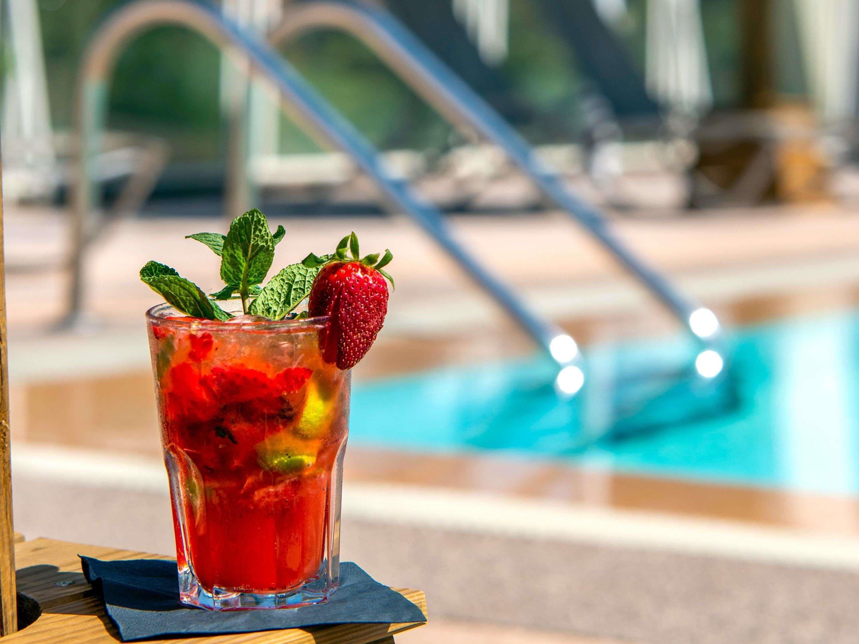 Un vaso de bebida roja con una fresa y una ramita de menta, sobre una superficie de madera con una piscina al fondo.