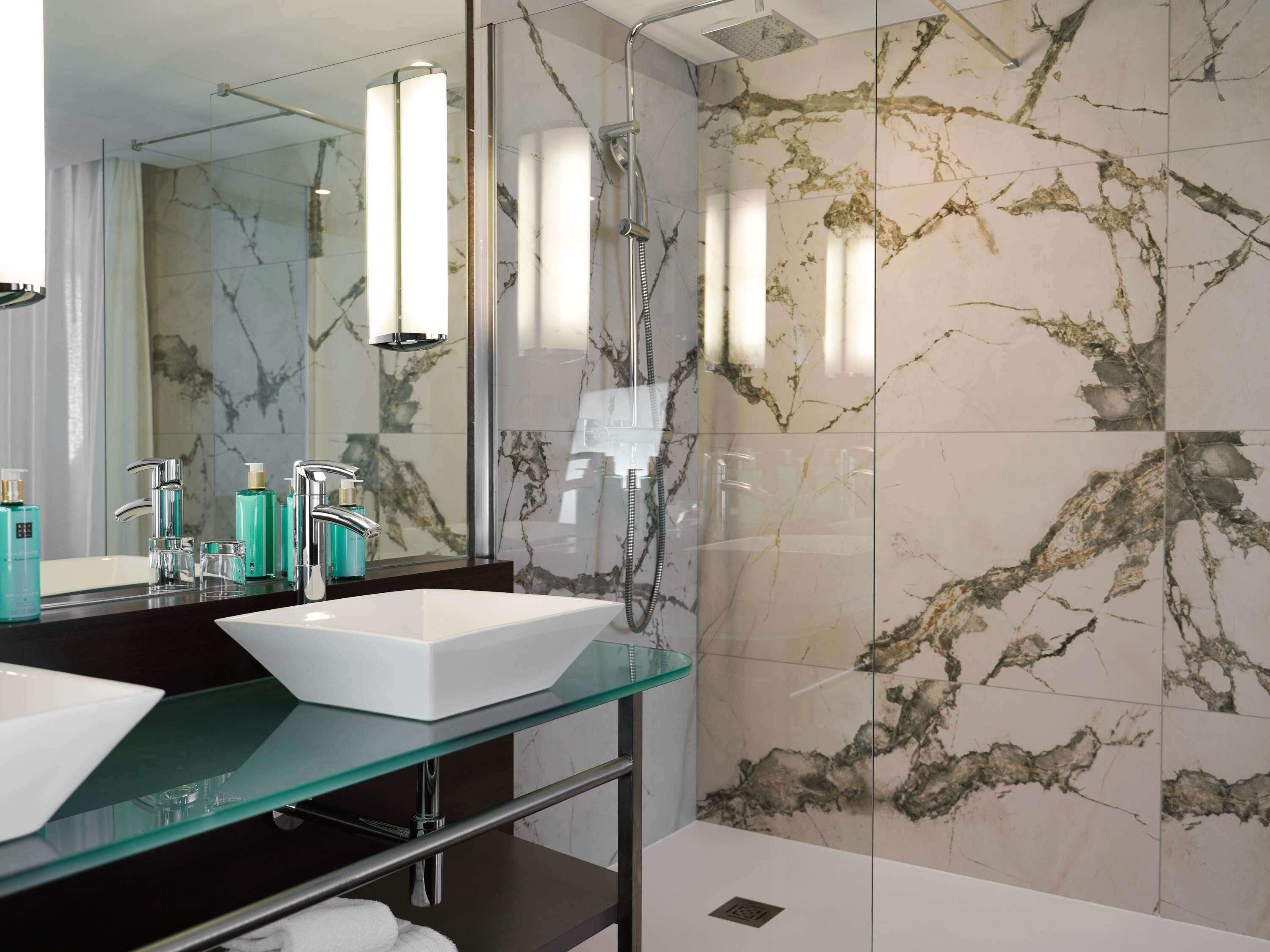 A modern bathroom with a glass vanity, two sinks, a shower, and marble walls.