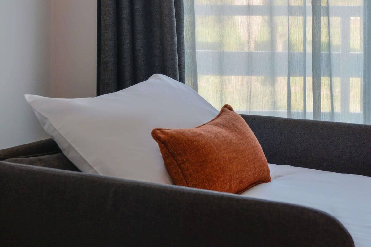 Un lit avec une literie blanche et un oreiller marron, un canapé de couleur foncée et une fenêtre donnant sur des arbres.