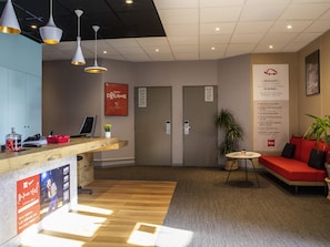 Een receptiezone met een houten toonbank, een computer, een plant, een rode bank en een muur met een bord.