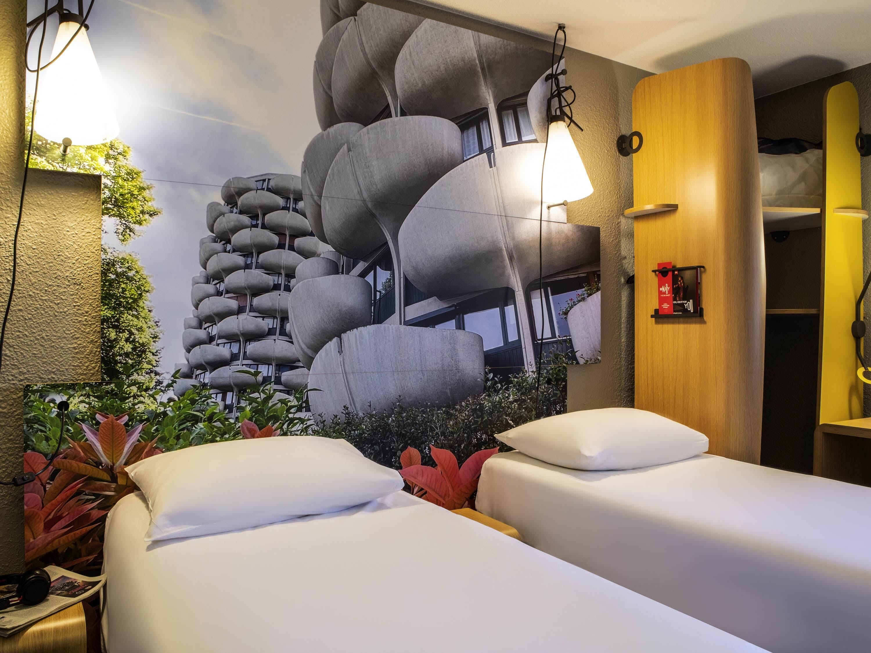 A hotel room with two beds, a wall mural, a wooden wardrobe, and a small plant.