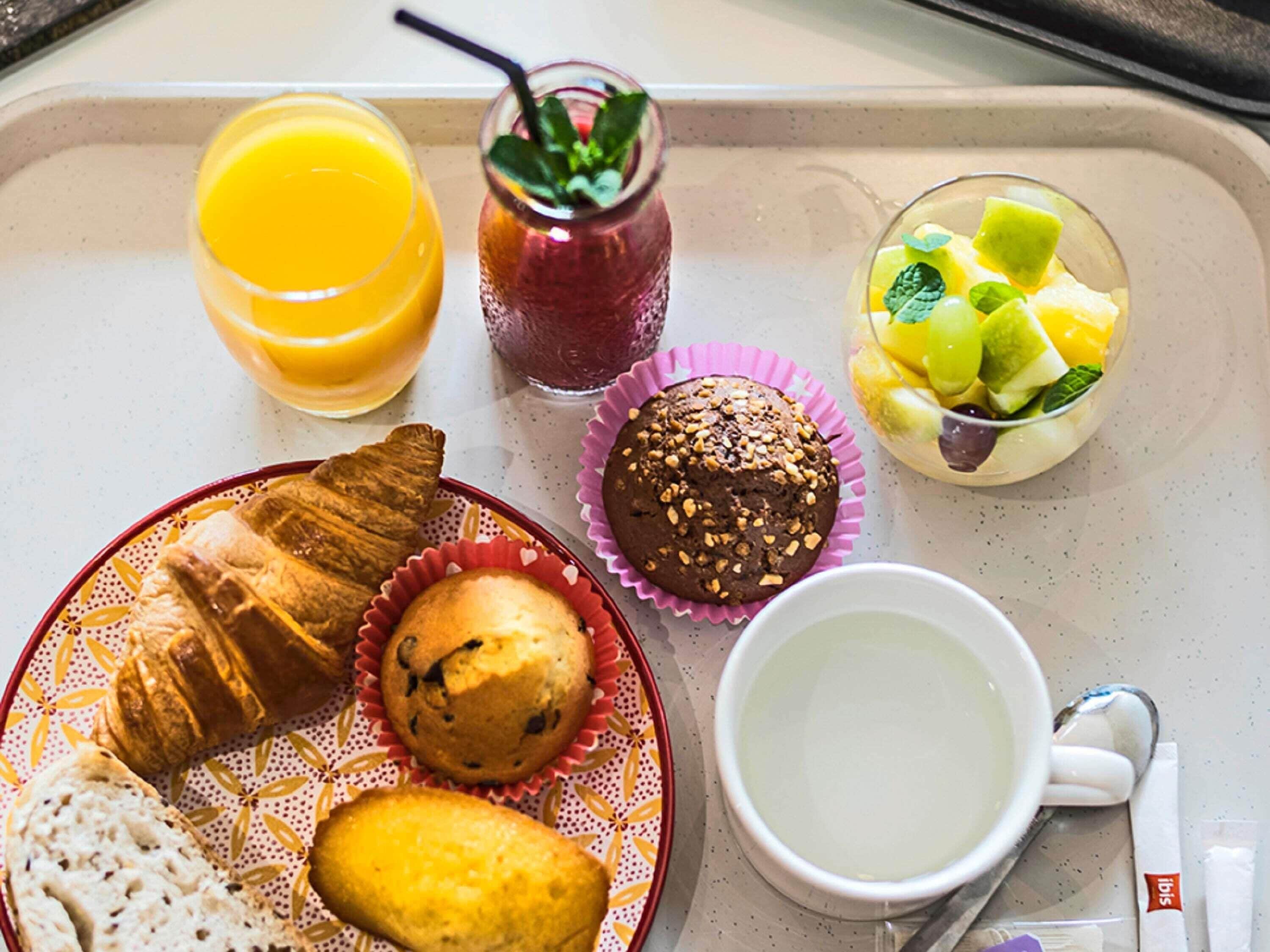 Bandeja de desayuno con croissants, muffins, un vaso de jugo de naranja, un bol de fruta y una taza de café.