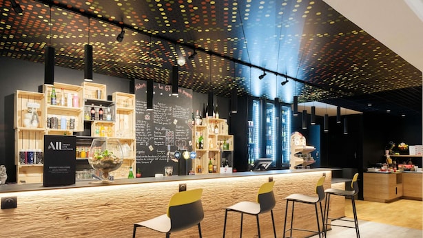 A bar area with a wooden counter, bar stools, and shelves with bottles. - ibis Strasbourg Sud La Vigie (Ostwald)