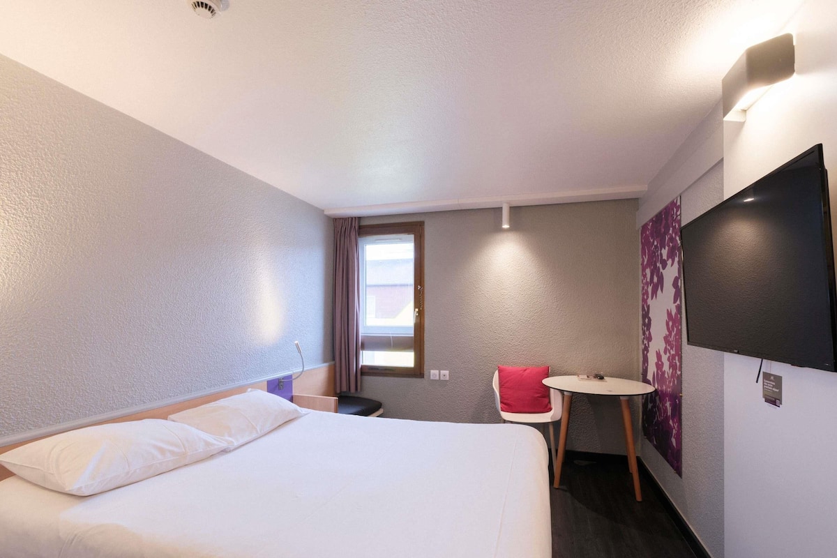 A hotel room with a bed, a small table, a chair, a TV, and a window.