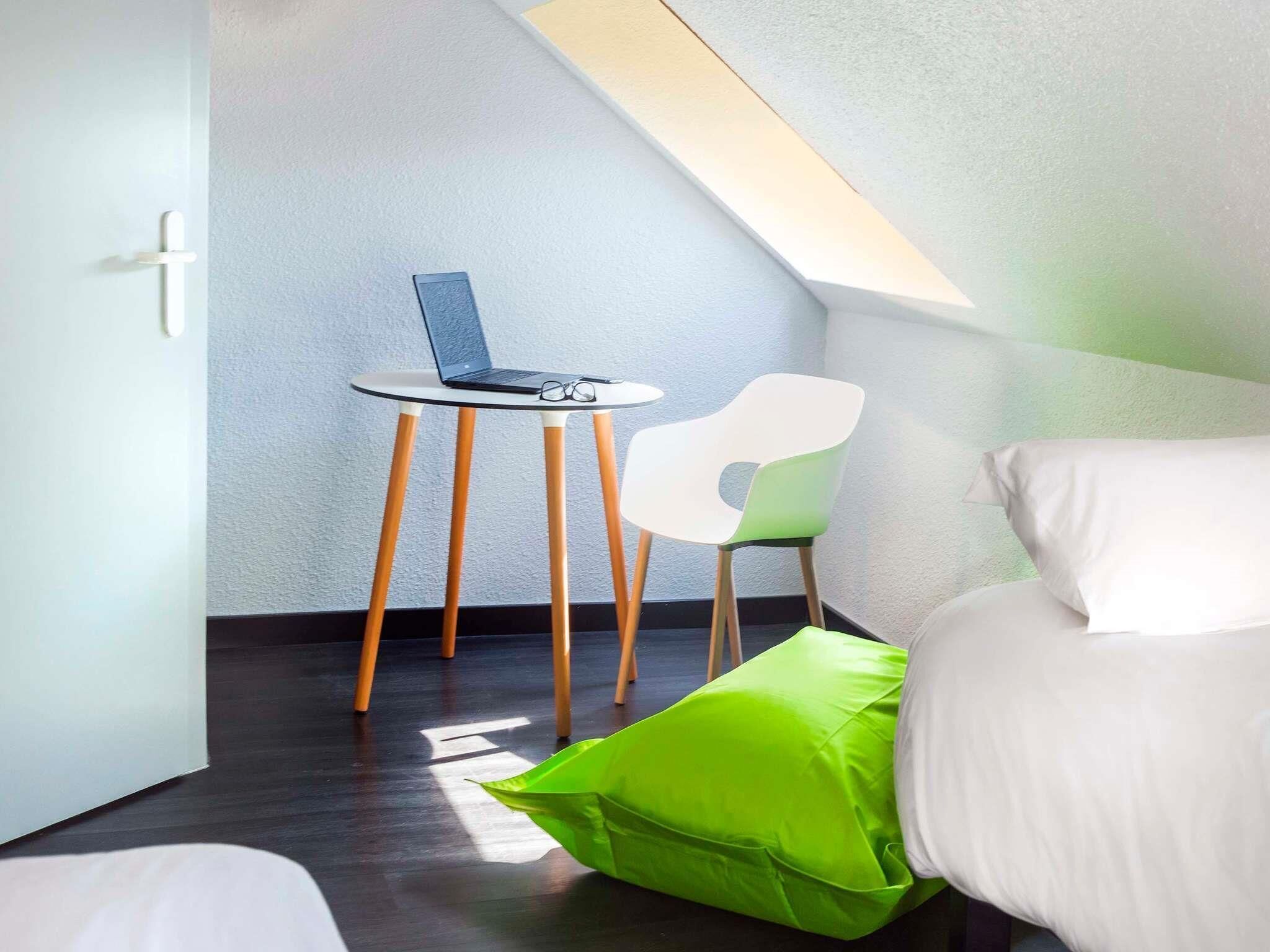 Una stanza piccola e moderna con soffitto spiovente, un tavolo rotondo con un portatile, una sedia verde e un letto con biancheria bianca.