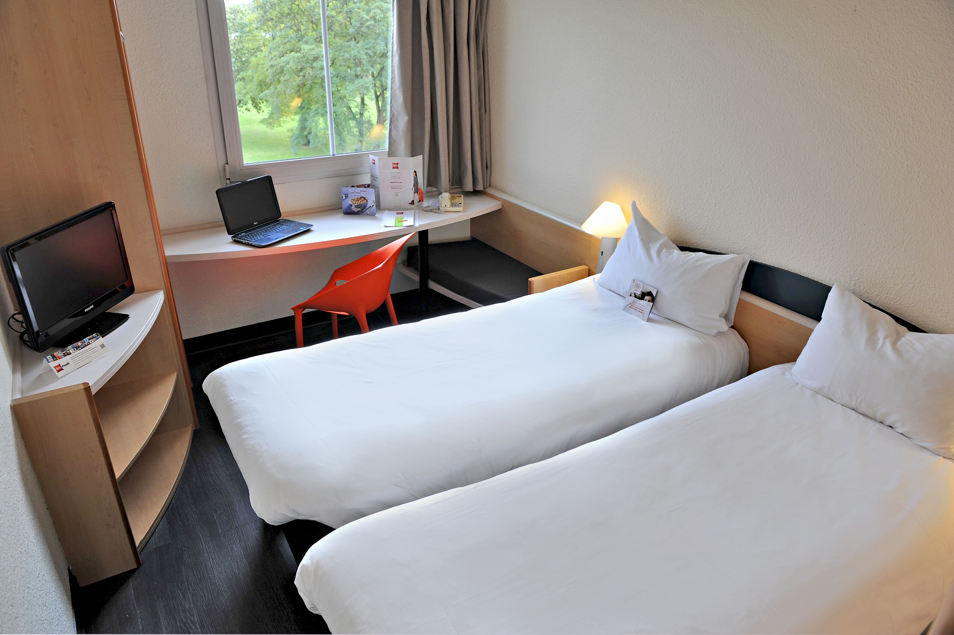 A hotel room with two beds, a desk with a laptop, a chair, and a window with curtains.