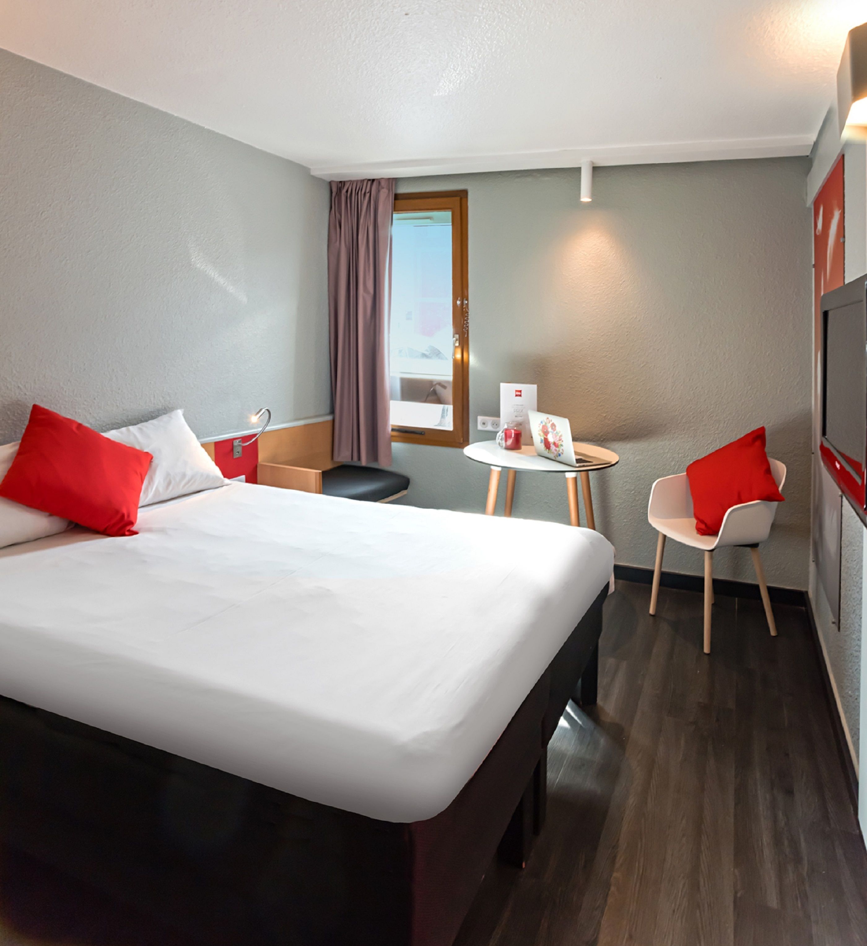 A hotel room with a large bed, a small table with a chair, a window with curtains, and a TV on the wall.